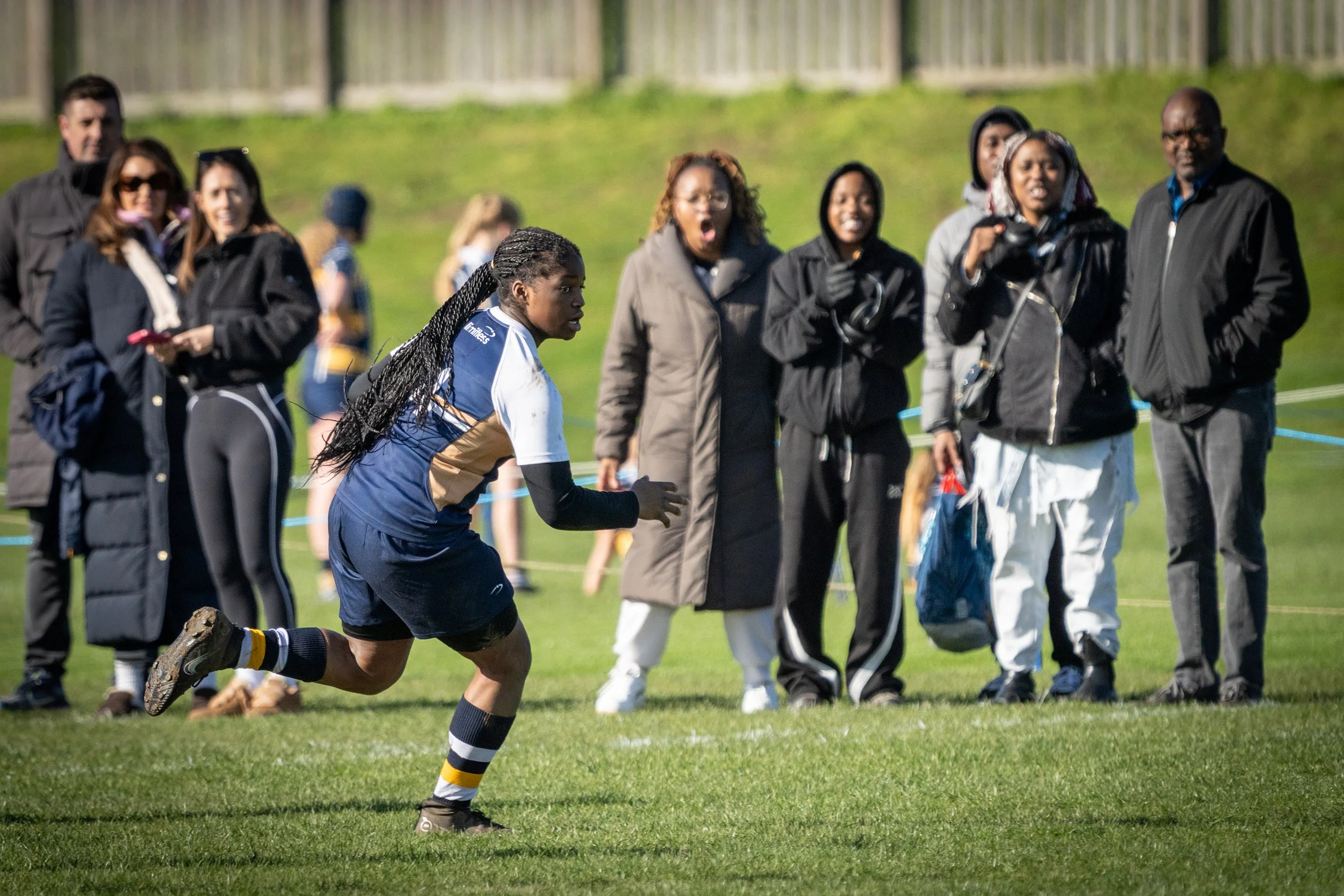 A female rugby player in a blue jersey running on a grassy field during a match, with a group of spectators watching behind her.