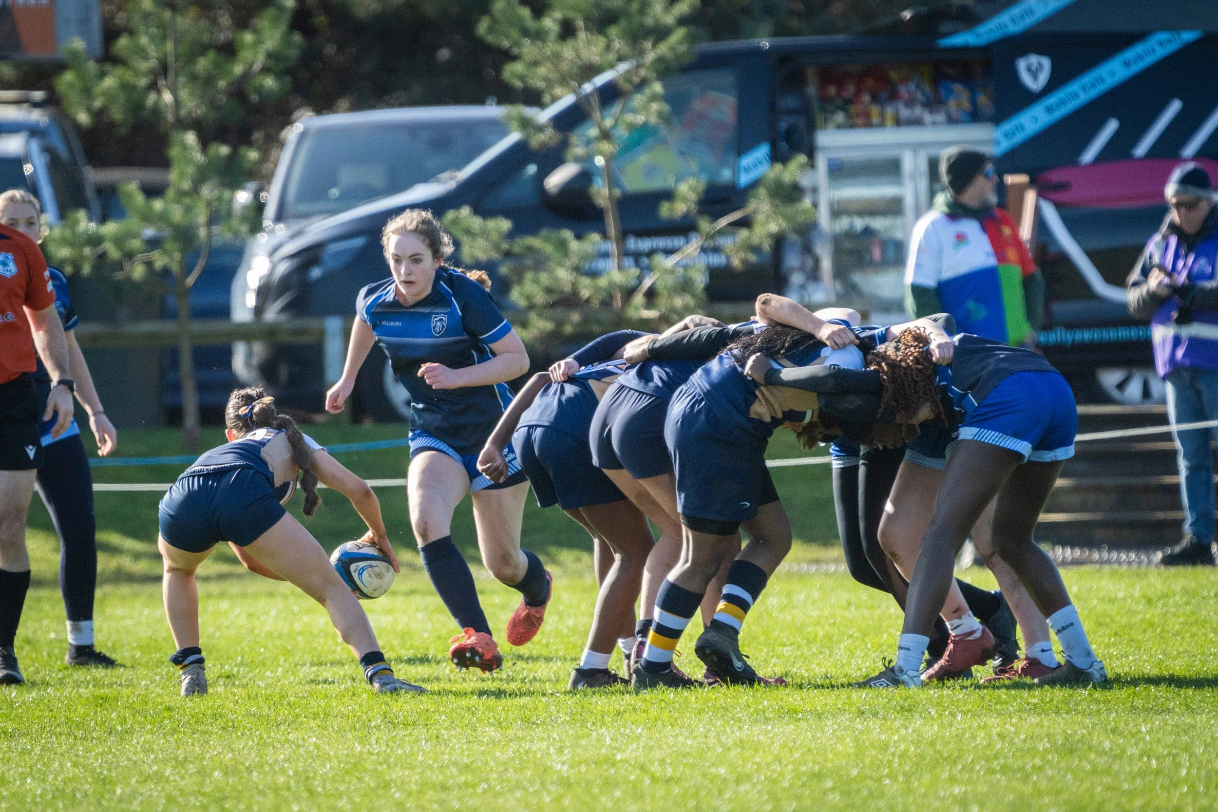 Women playing rugby on a grassy field, some leaning forward and others standing, with vehicles and trees in the background.