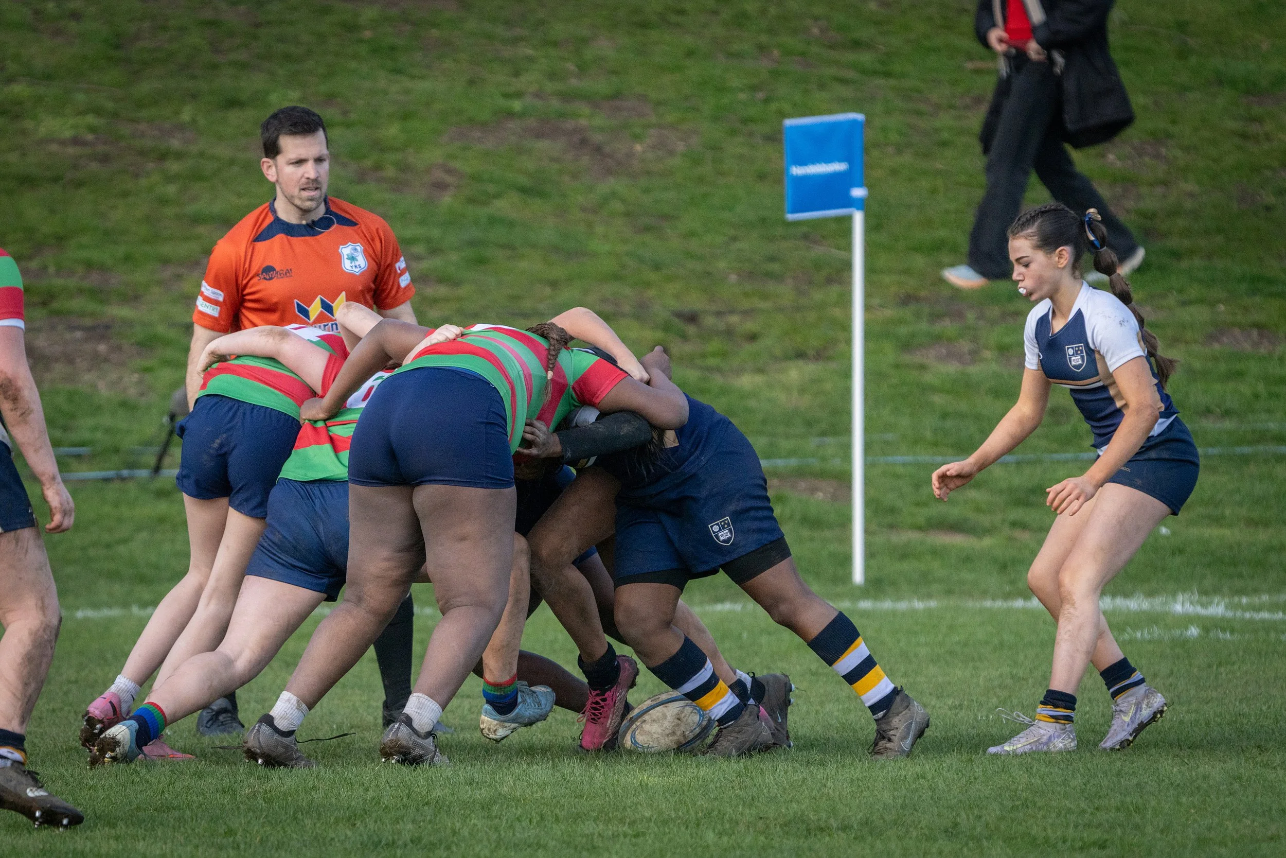 A women's rugby match with players engaged in a scrum on a grassy field, referees, and a person walking nearby.