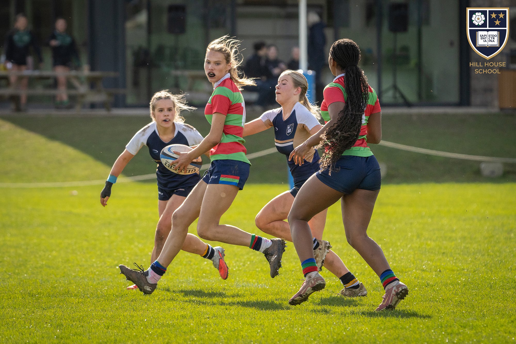 Young girls playing rugby on a grassy field, with a girl in a white and blue uniform holding a rugby ball and other girls in colorful striped shirts and shorts running around her during daylight.