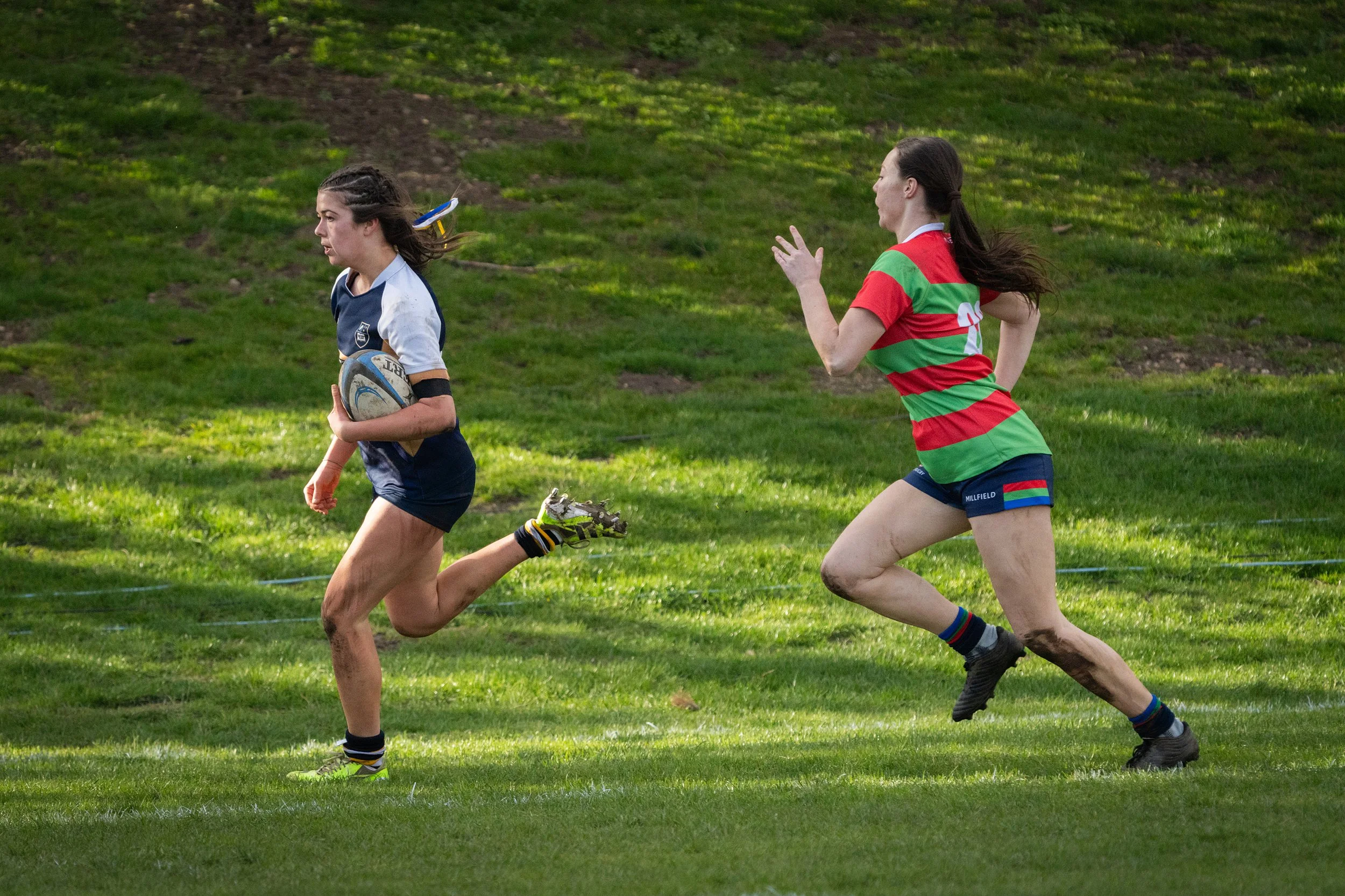 Two women playing rugby on grass field with one holding a rugby ball and both running swiftly.