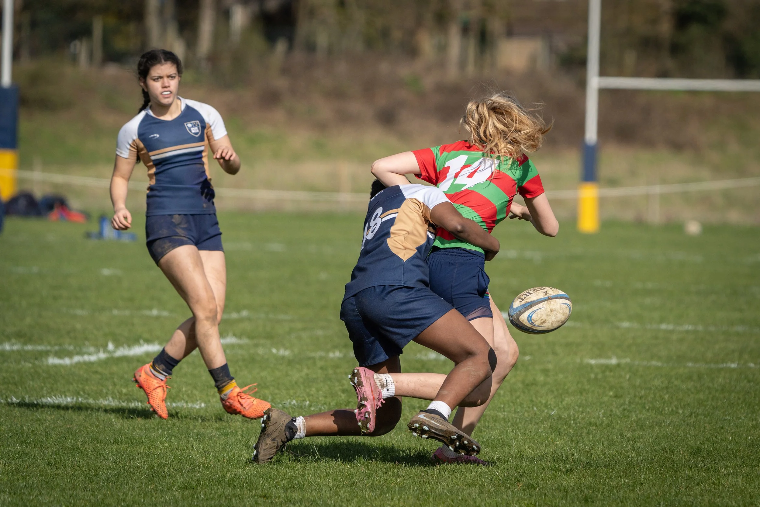 Girls playing rugby on a grassy field, one girl in a red and green uniform is tackled by another in a blue and gold uniform, with a third girl watching nearby.