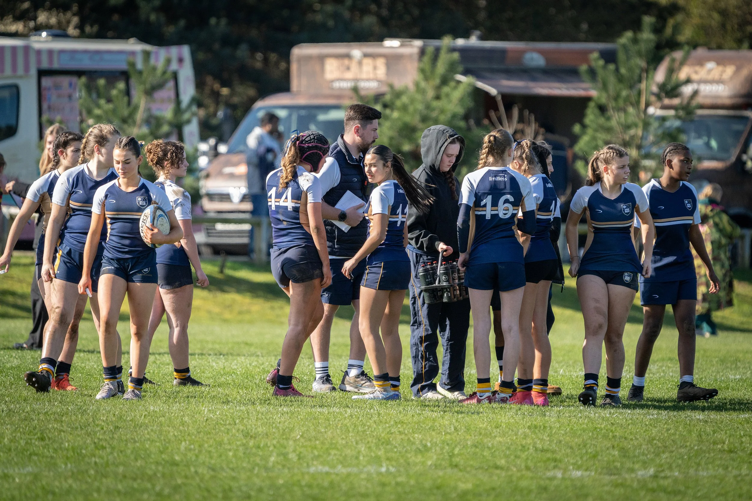 Group of female rugby players wearing blue and white uniforms walking on a grassy field with trees and food trucks in the background.