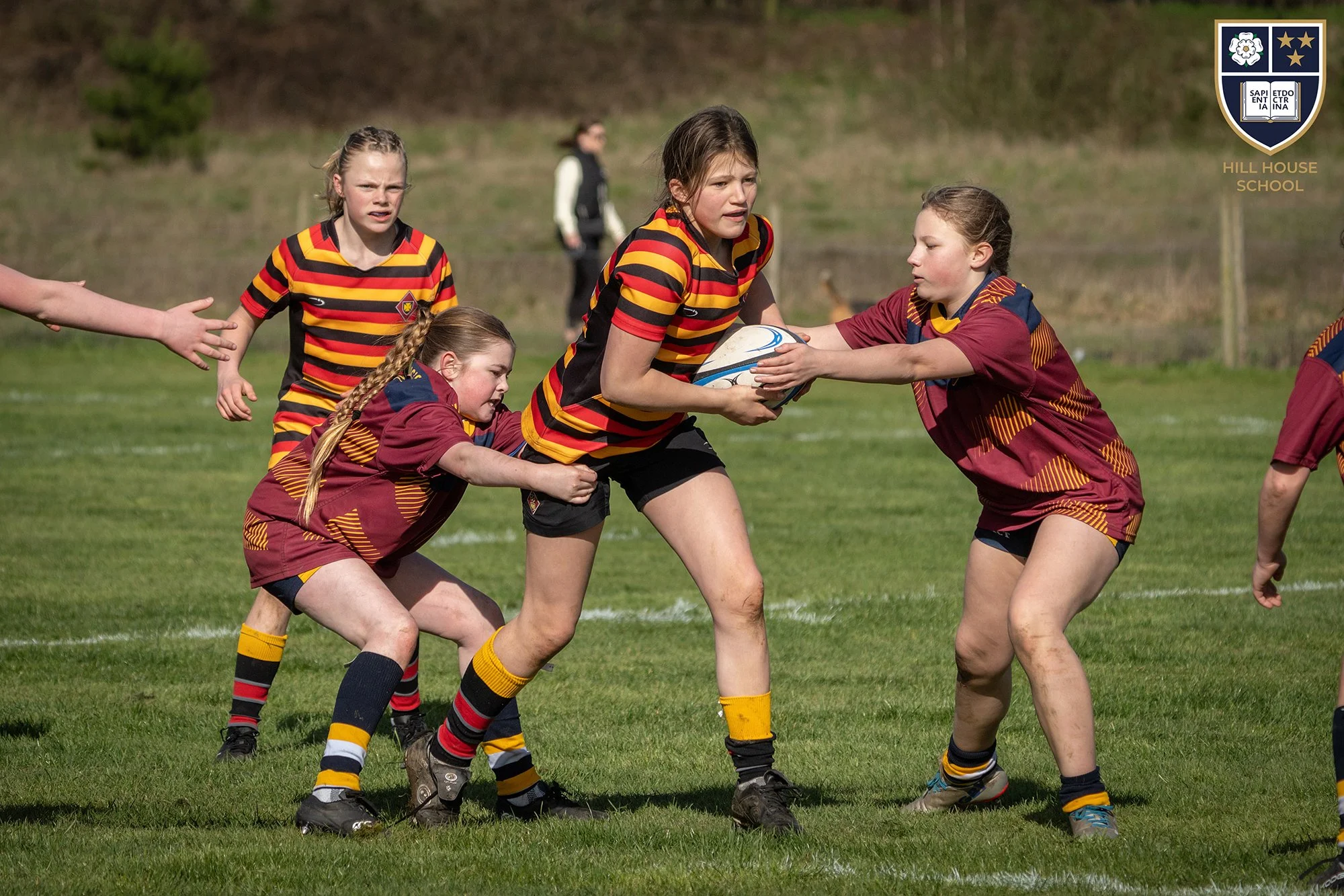 Young girls playing rugby on a grassy field, with some girls wearing yellow, red, and black striped jerseys and others in maroon jerseys, engaging in a game with one girl holding a rugby ball as others try to stop her.