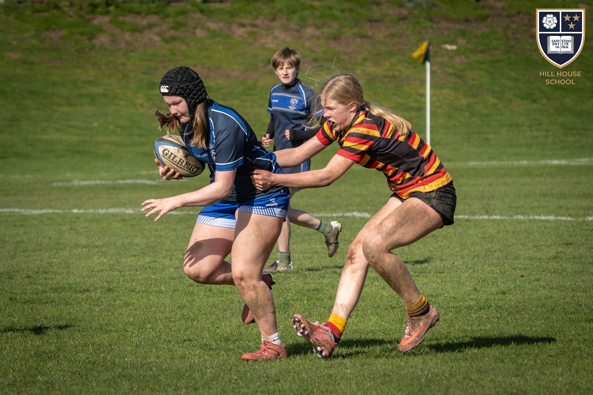 Three children playing rugby on a grassy field. One girl with a rugby helmet is holding a rugby ball while another girl with a striped rugby shirt is attempting to tackle her. A boy in a blue rugby uniform runs in the background.