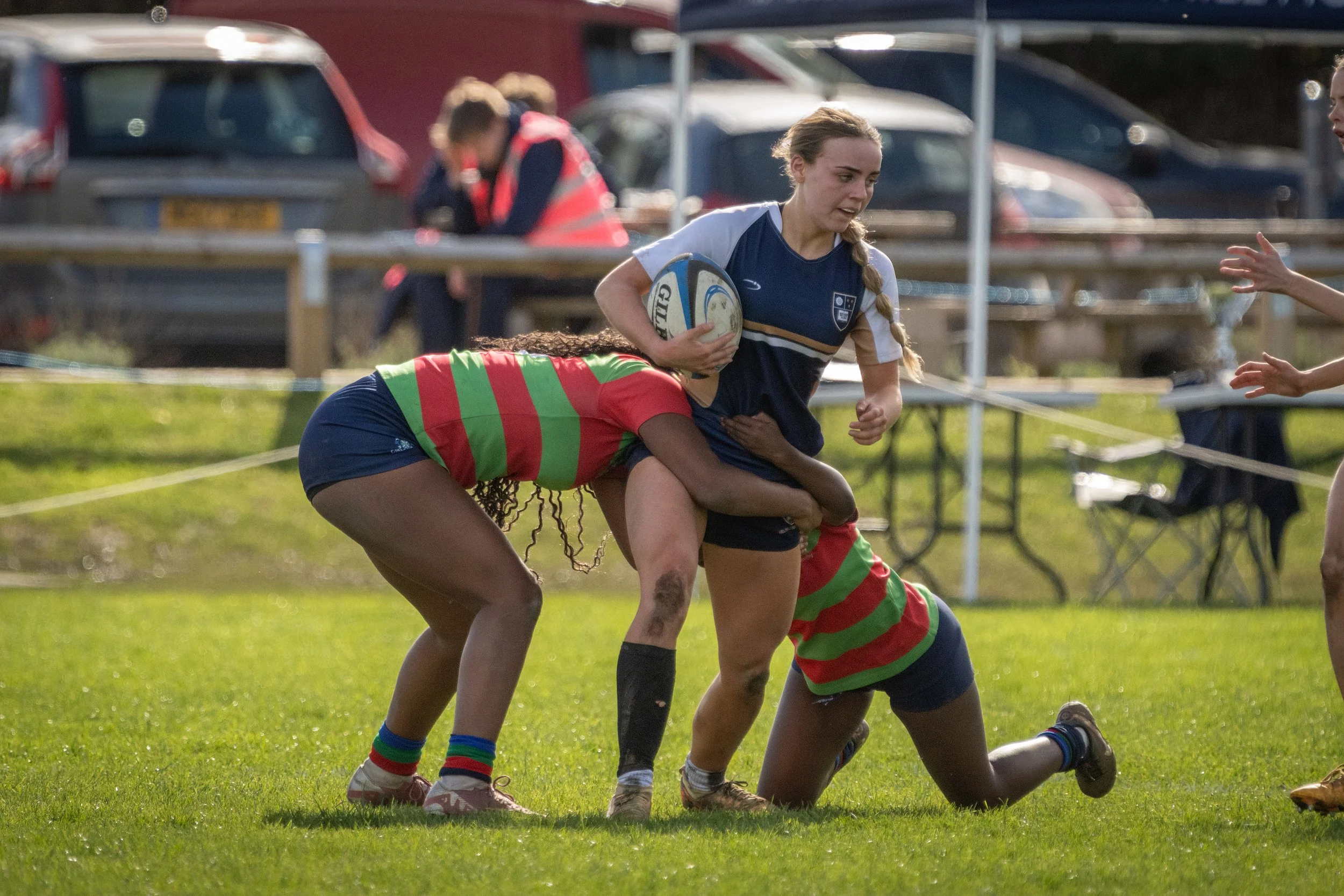 Women playing rugby on a grassy field, with one holding a rugby ball and others engaged in a tackle.