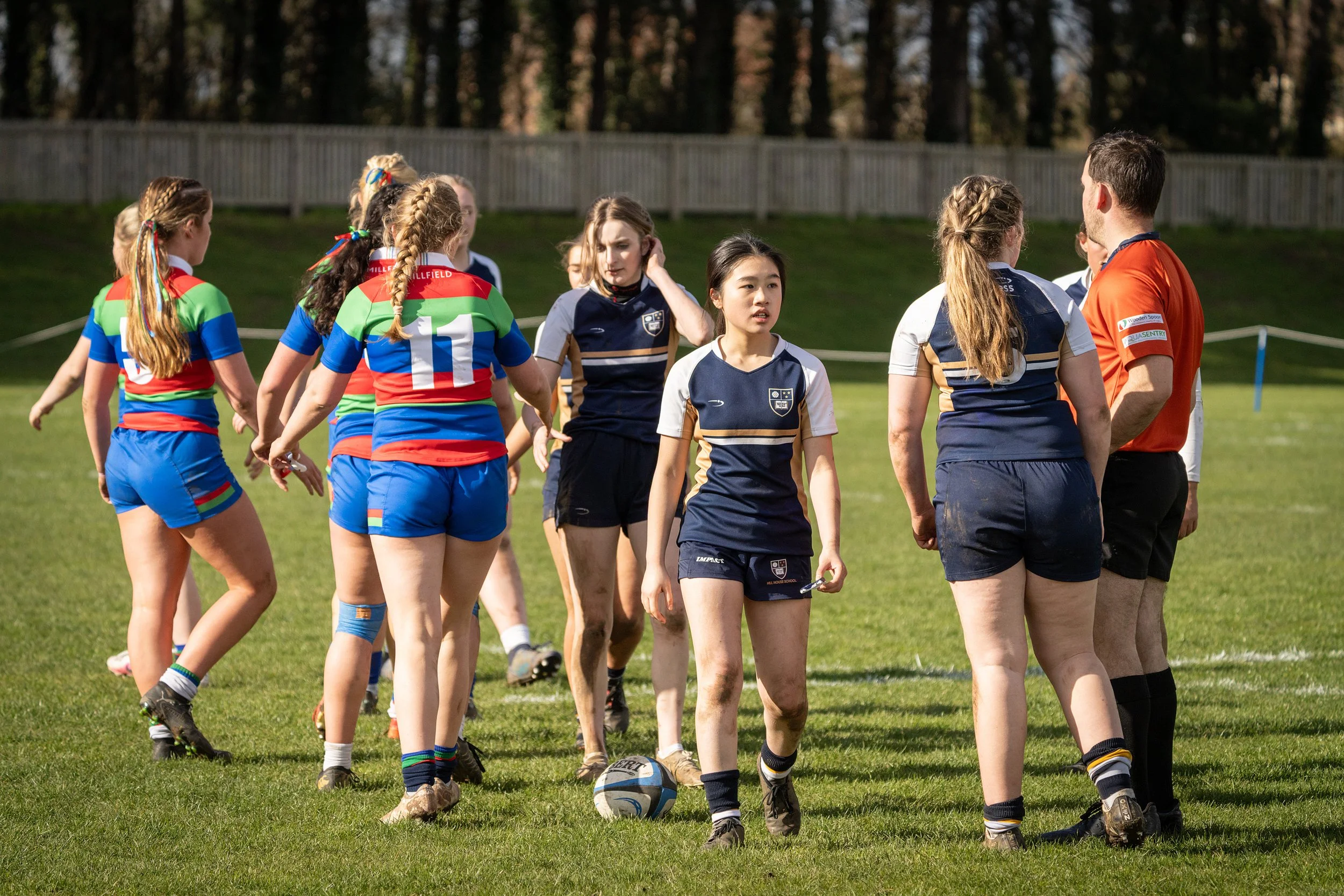 A group of young female rugby players and a male coach are gathered on a grassy field during daylight, preparing for or during a match, with a wooden fence and trees in the background.
