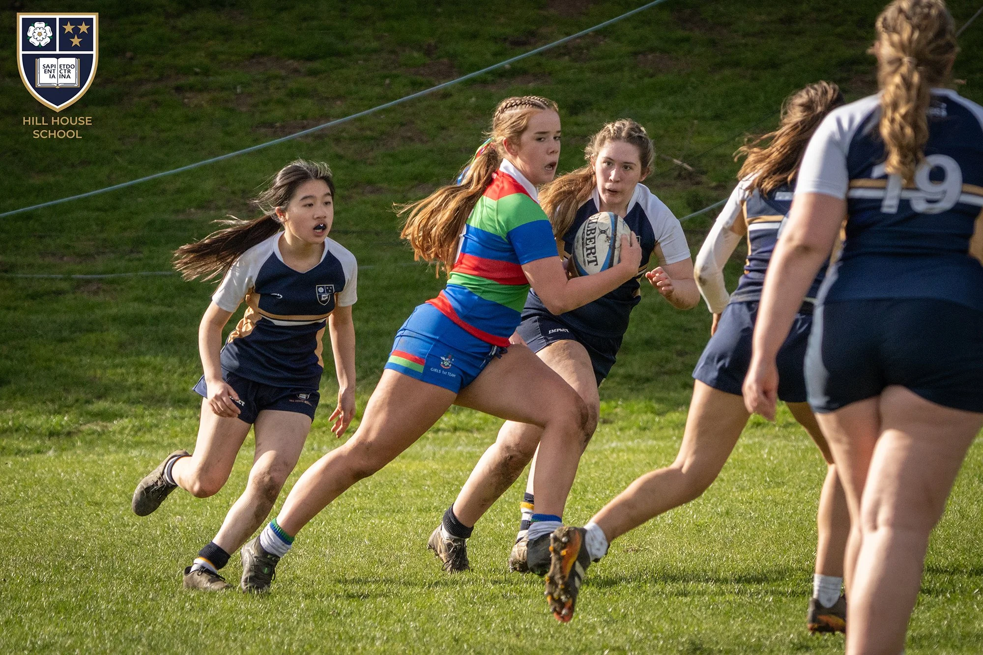 Girls rugby game on a grassy field at Hill House School, one girl in a rainbow striped uniform holding a rugby ball, other girls in navy and white uniforms running.