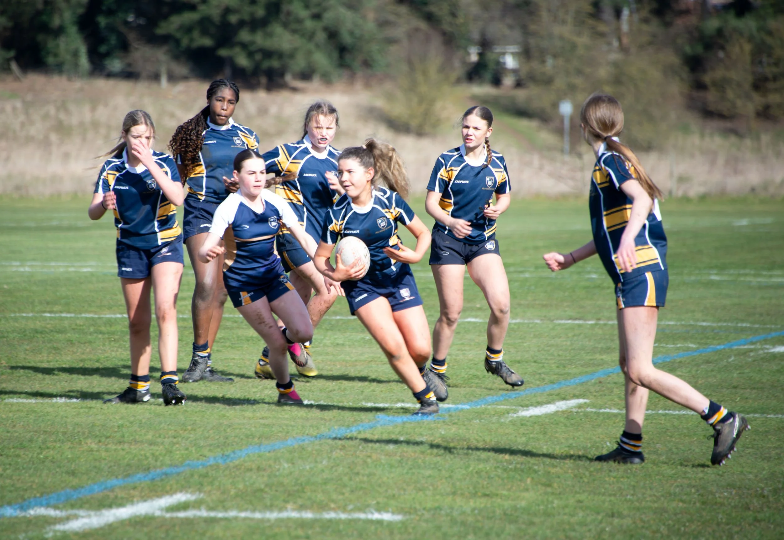Girls playing rugby on a grassy field during daytime, wearing blue and yellow uniforms.