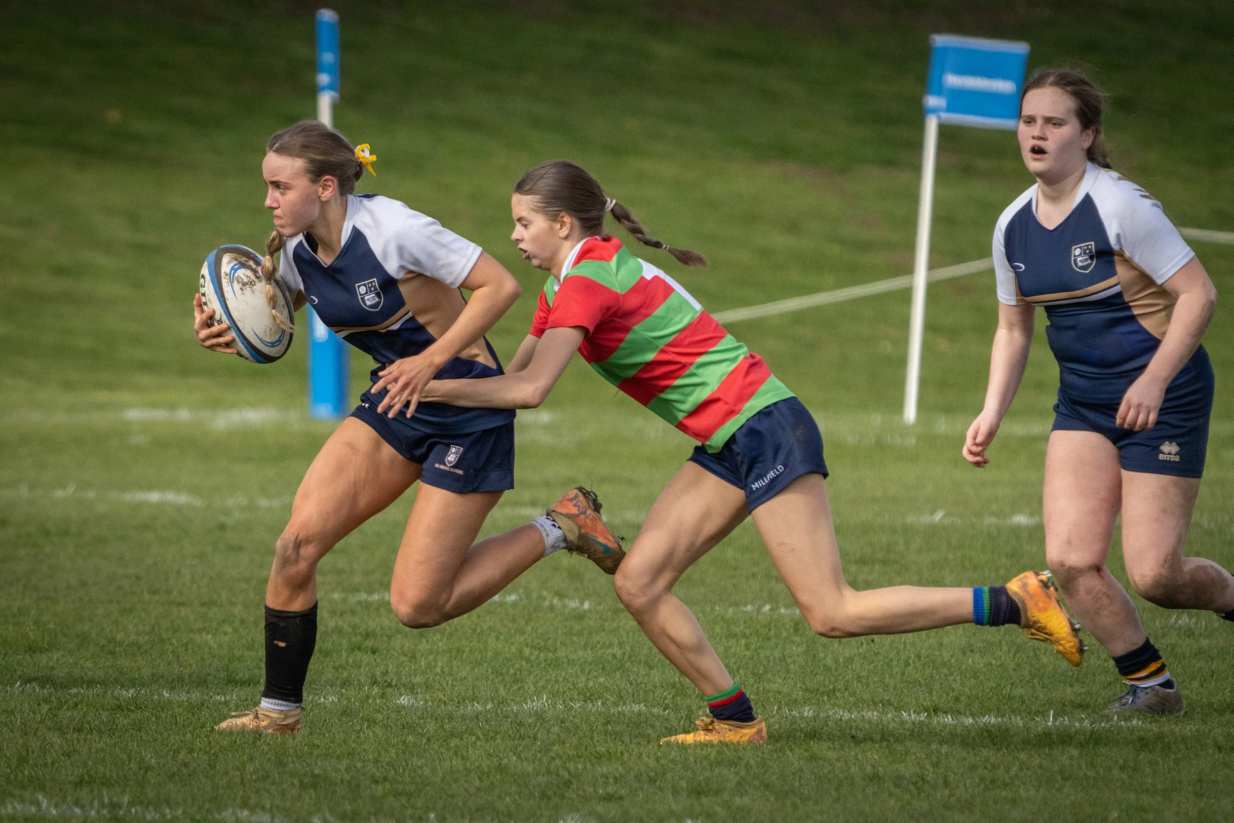 Girls playing rugby on a grassy field, wearing sports uniforms with one girl running holding a rugby ball, two others chasing behind, during a game.