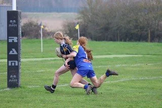 Two female rugby players competing for the ball on a grassy field, with one in black and yellow and the other in blue, during a match.