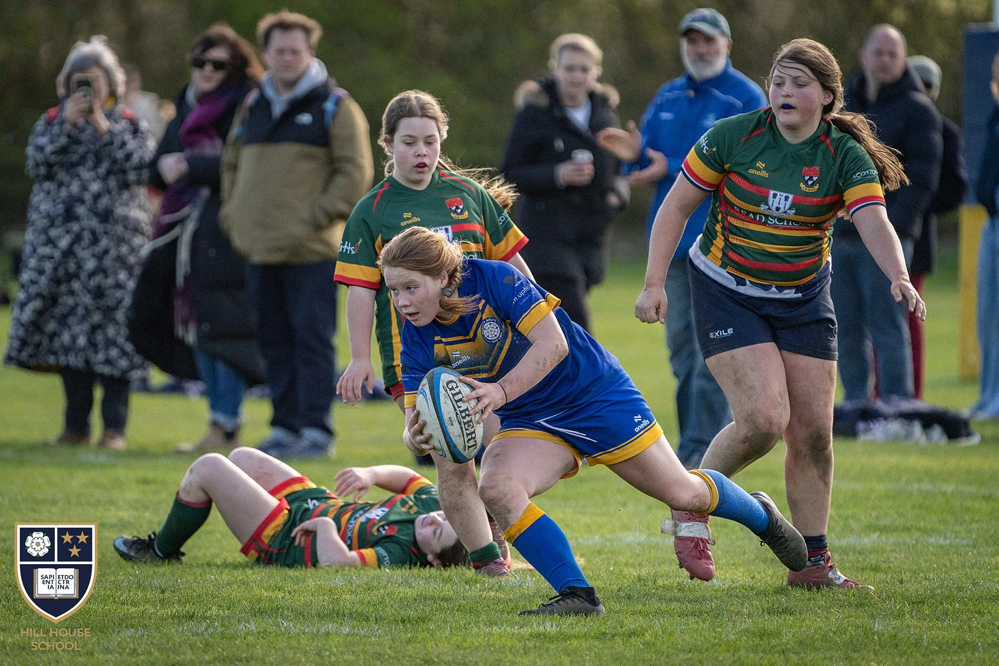 Young girls playing rugby on a grassy field with spectators watching in the background. One girl in a green and yellow uniform is tackling another girl in a blue uniform who is holding a rugby ball. The scene captures action and teamwork during a rug