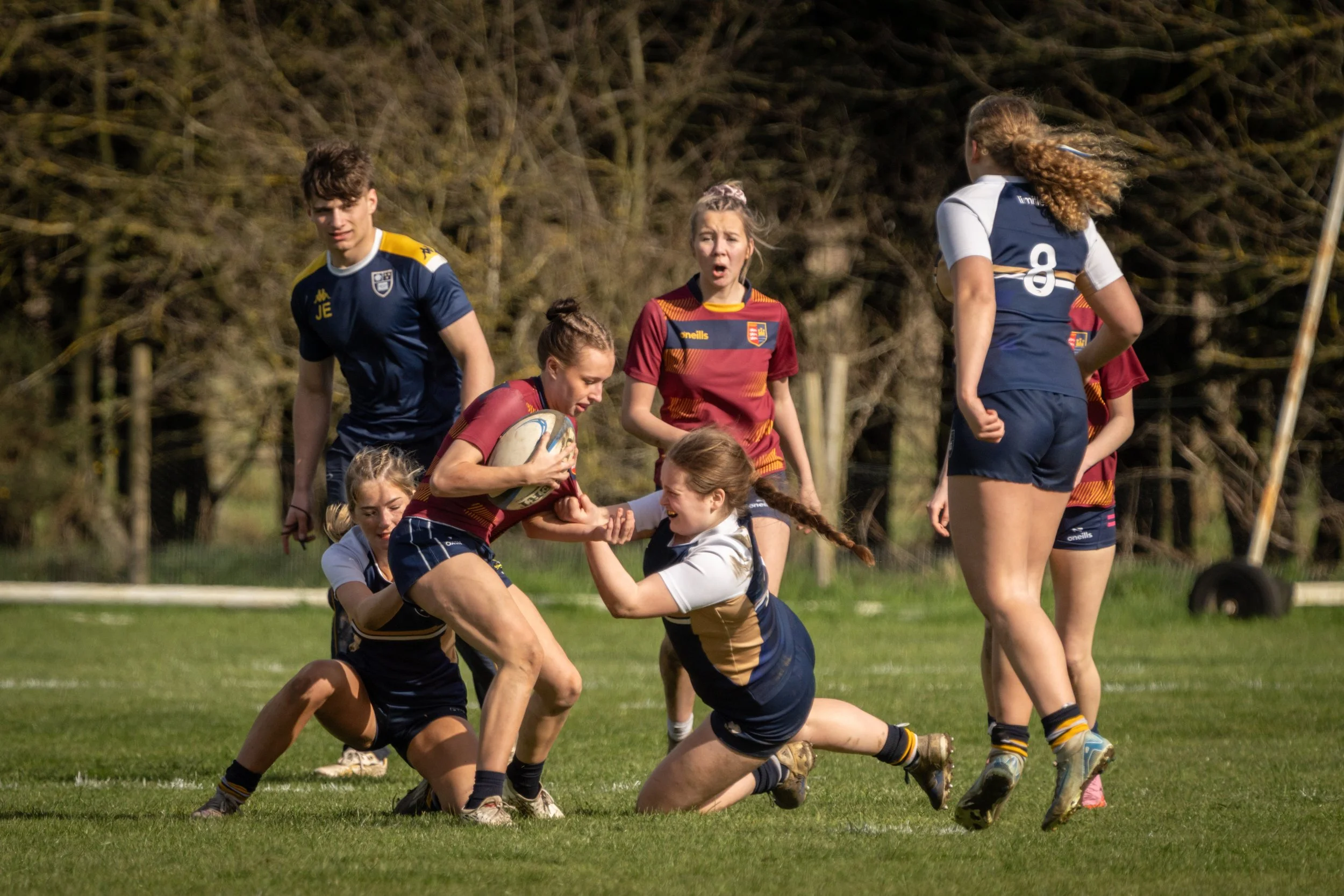 Young women playing rugby on a grassy field, some tackling and others standing nearby, with trees in the background.