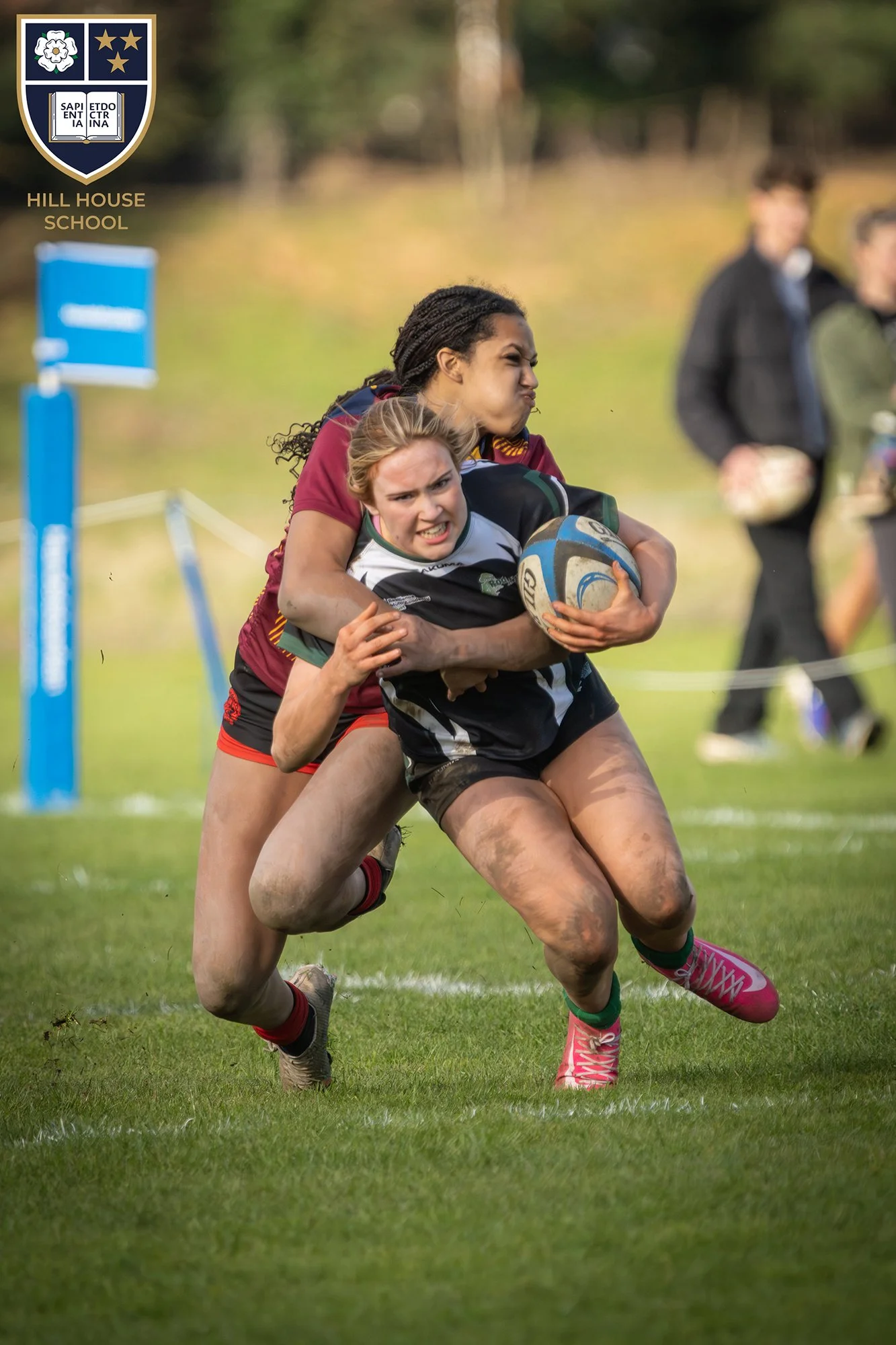 Two women playing rugby on a grass field, one woman holding a rugby ball, wearing sportswear and mud-stained legs, with streaks of mud and grass. In the background, there are spectators, and a Hill House School logo is in the top left corner.