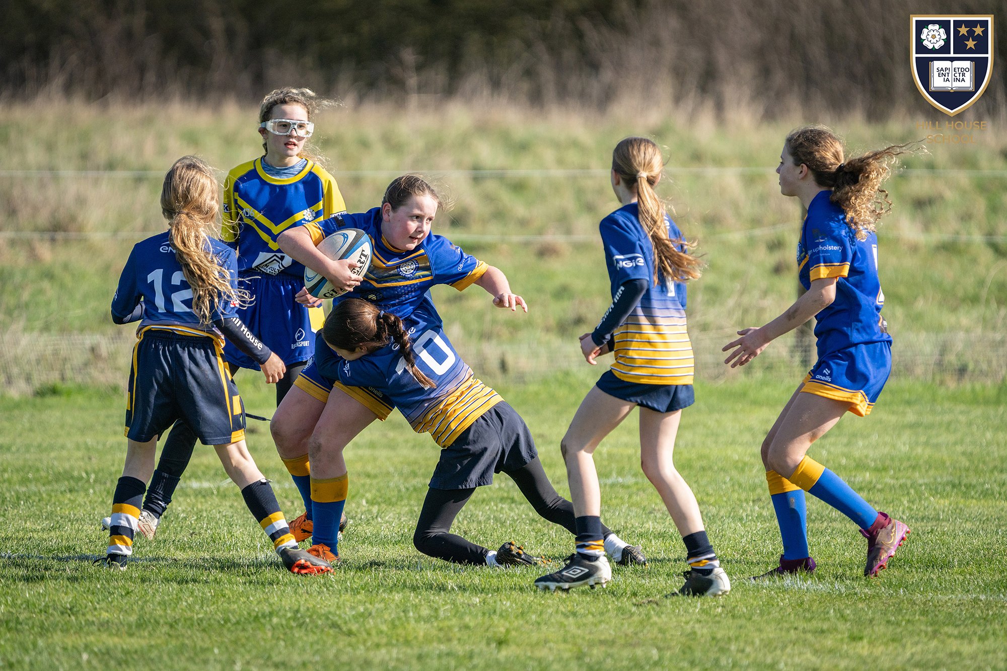 Young girls playing rugby on a grassy field, wearing blue and yellow team uniforms, with some girls engaging in a tackle while others watch or approach, and a school crest with a flower, stars, and a classical book logo in the top right corner.