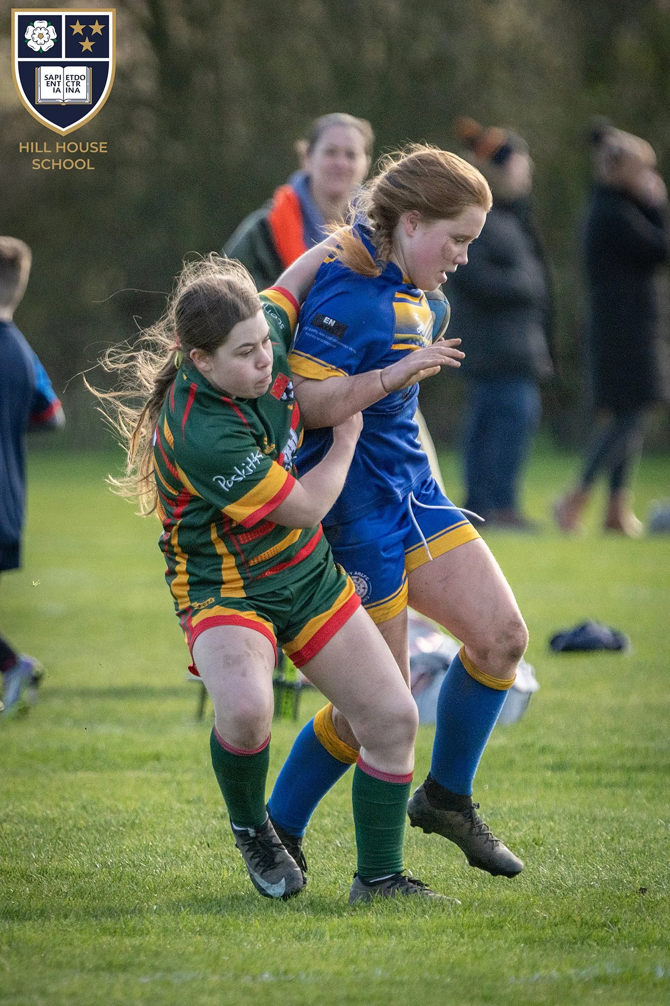 Two female rugby players, one in a green and red uniform and the other in a blue and yellow uniform, are engaged in a match, with the player in green attempting to tackle the player in blue.