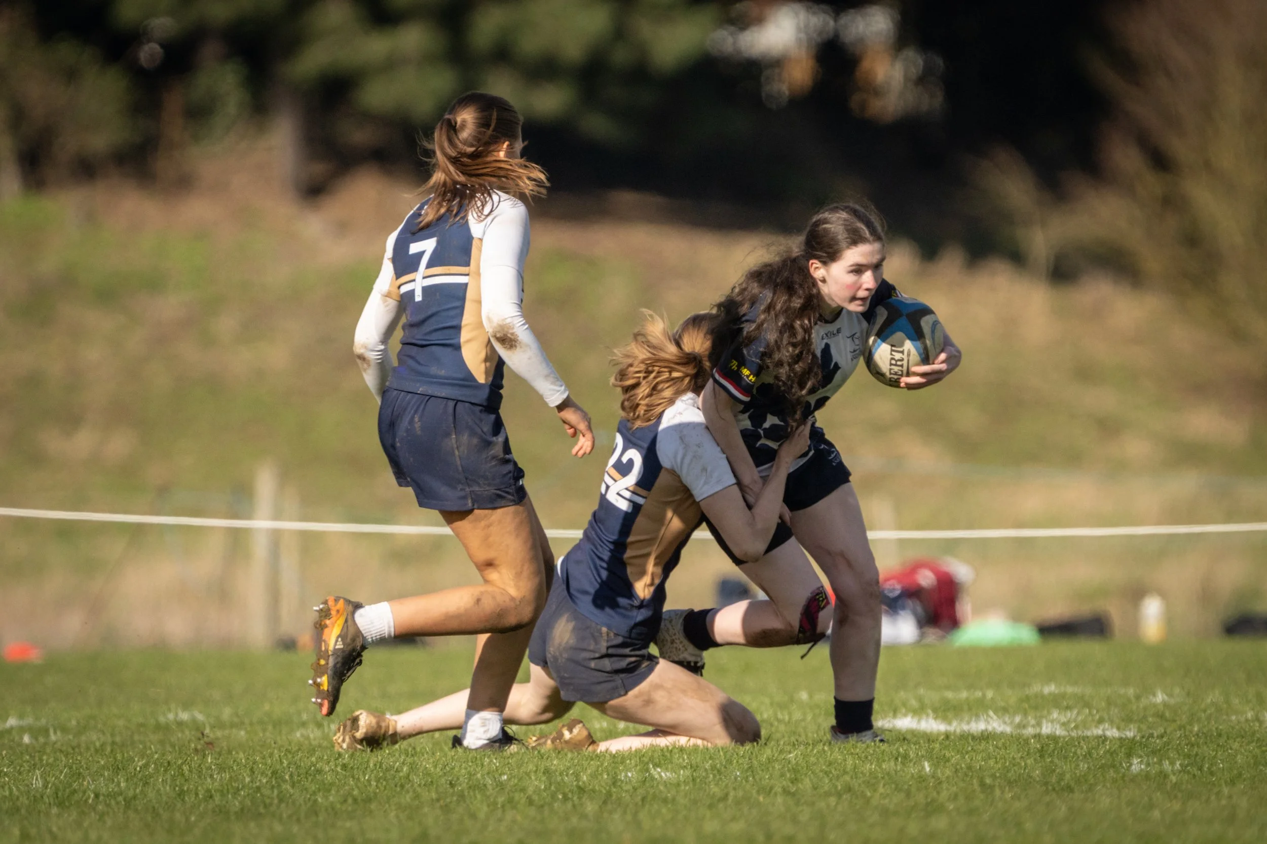 Women playing rugby on the field, with one woman holding a rugby ball and other women tackling in the game.