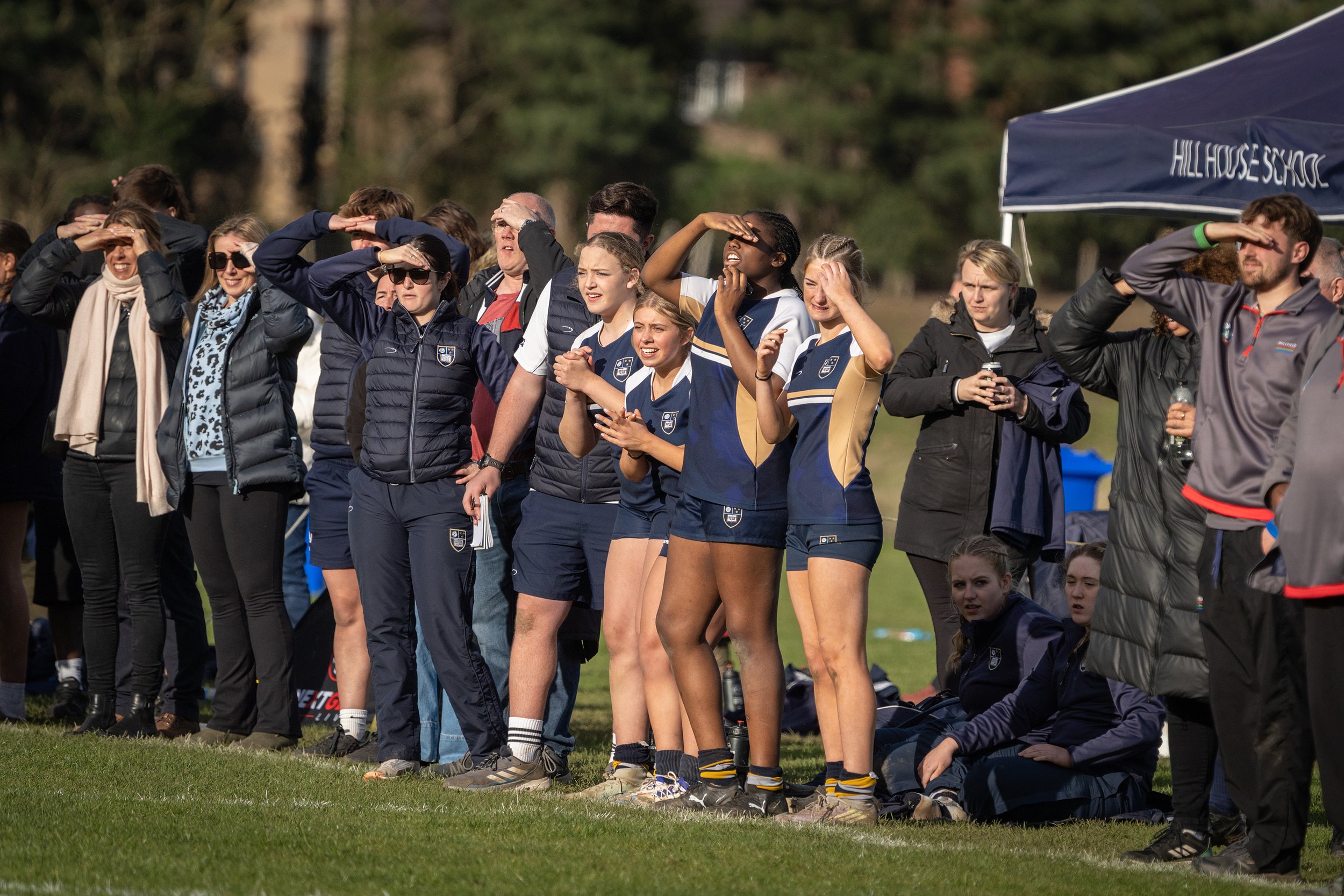 A group of female and male athletes and spectators standing outdoors, saluting during a sports event, with some wearing sports uniforms, standing near a blue tent with 'HILLHOUSE SCHOOL' written on it, in a grassy field with trees in the background.