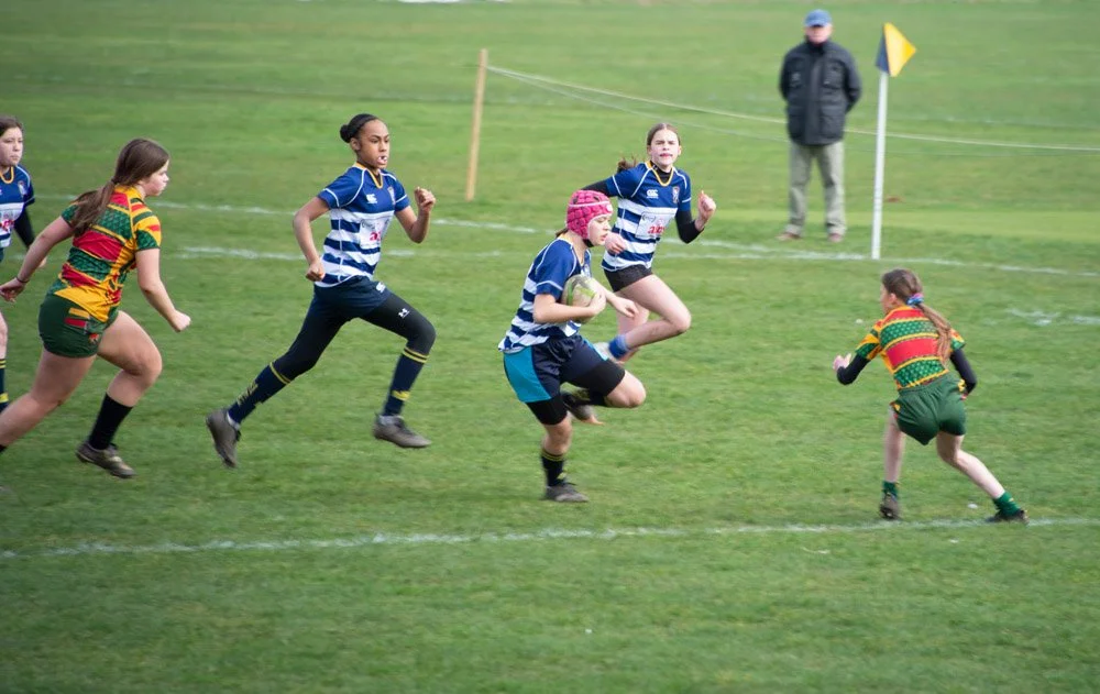 Children playing rugby on a grassy field, with players in blue and green uniforms, some running and one holding a rugby ball, and an adult standing in the background.