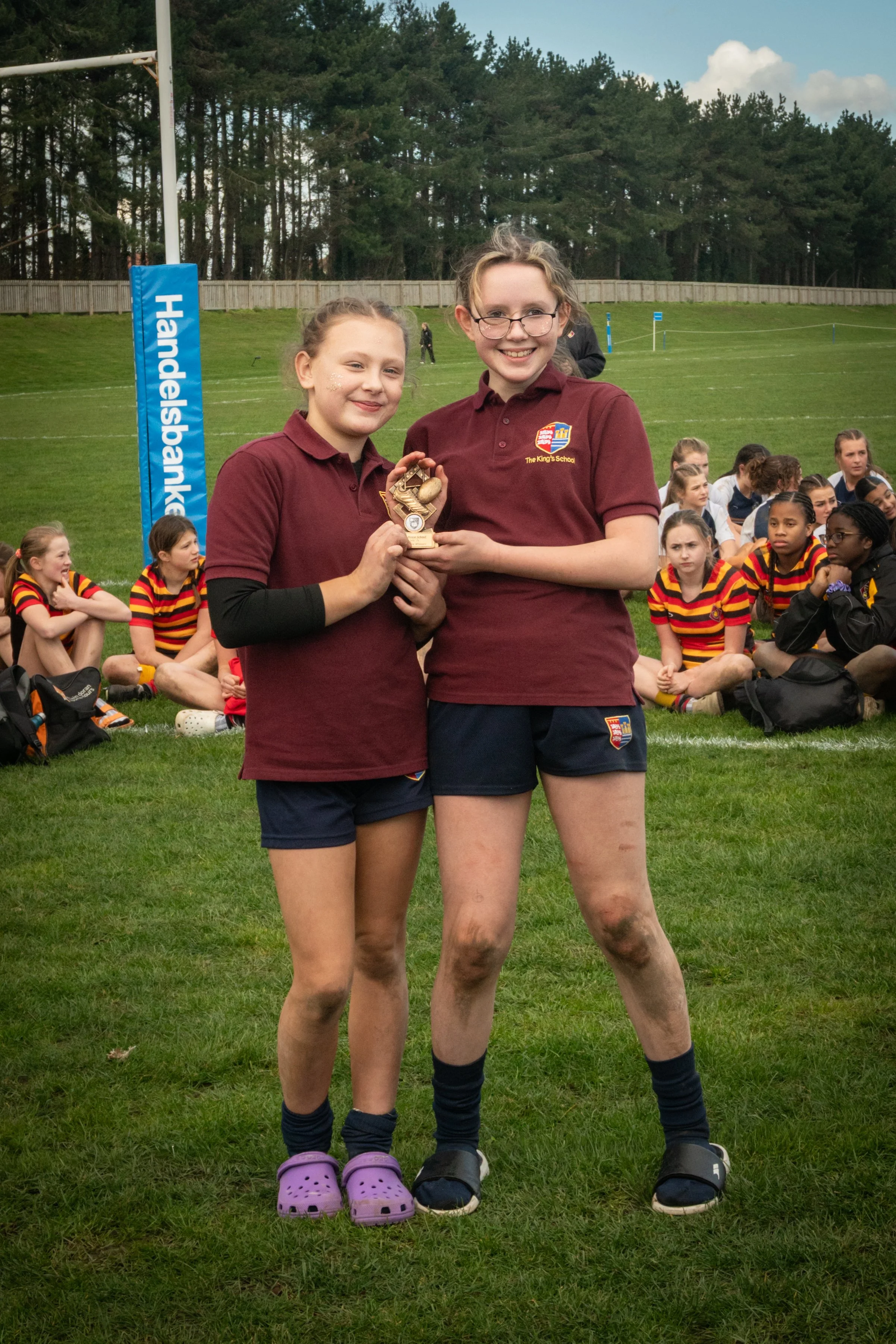 Two young girls in maroon sports uniforms holding a small wooden trophy on a grassy field, with a group of children seated behind them, some wearing striped shirts, and a rugby goalpost with a blue padding and the word 'Handlebanken' on it. Trees and a wooden fence are in the background.