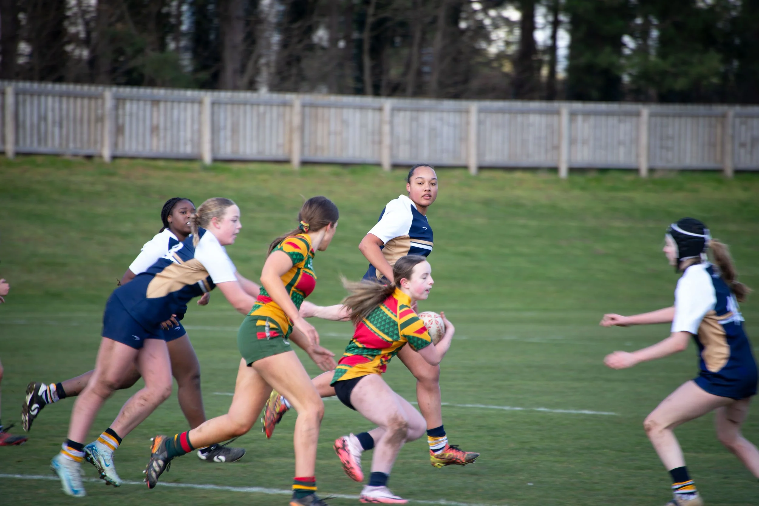 Young girls playing rugby on a grassy field, wearing colorful and sports uniforms, with a wooden fence and trees in the background.