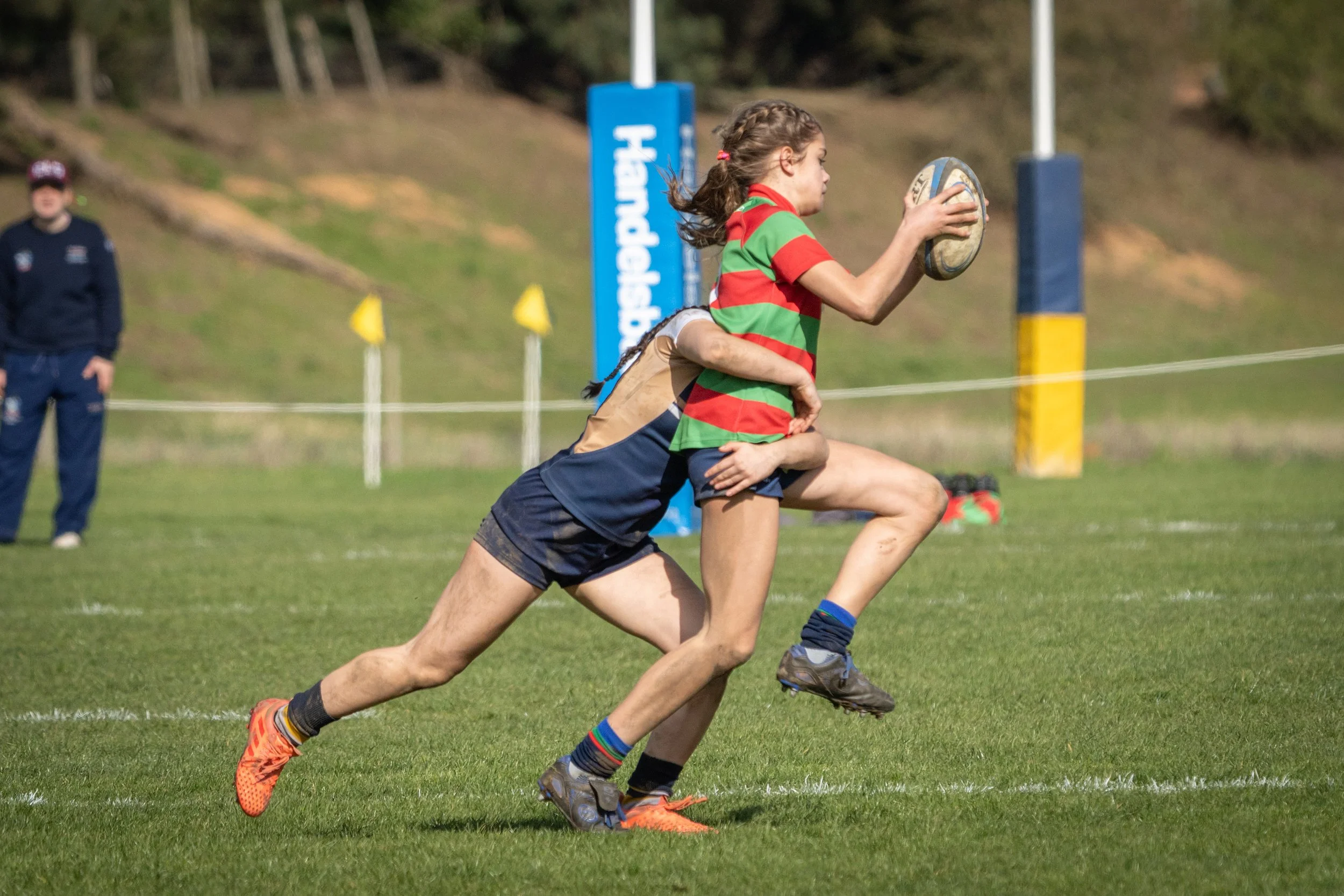 A young girl in a green and red striped shirt and shorts is running on a rugby field while holding a rugby ball. She is in mid-stride with her left leg extended forward and her right arm bent holding the ball close to her chest. In the background, th