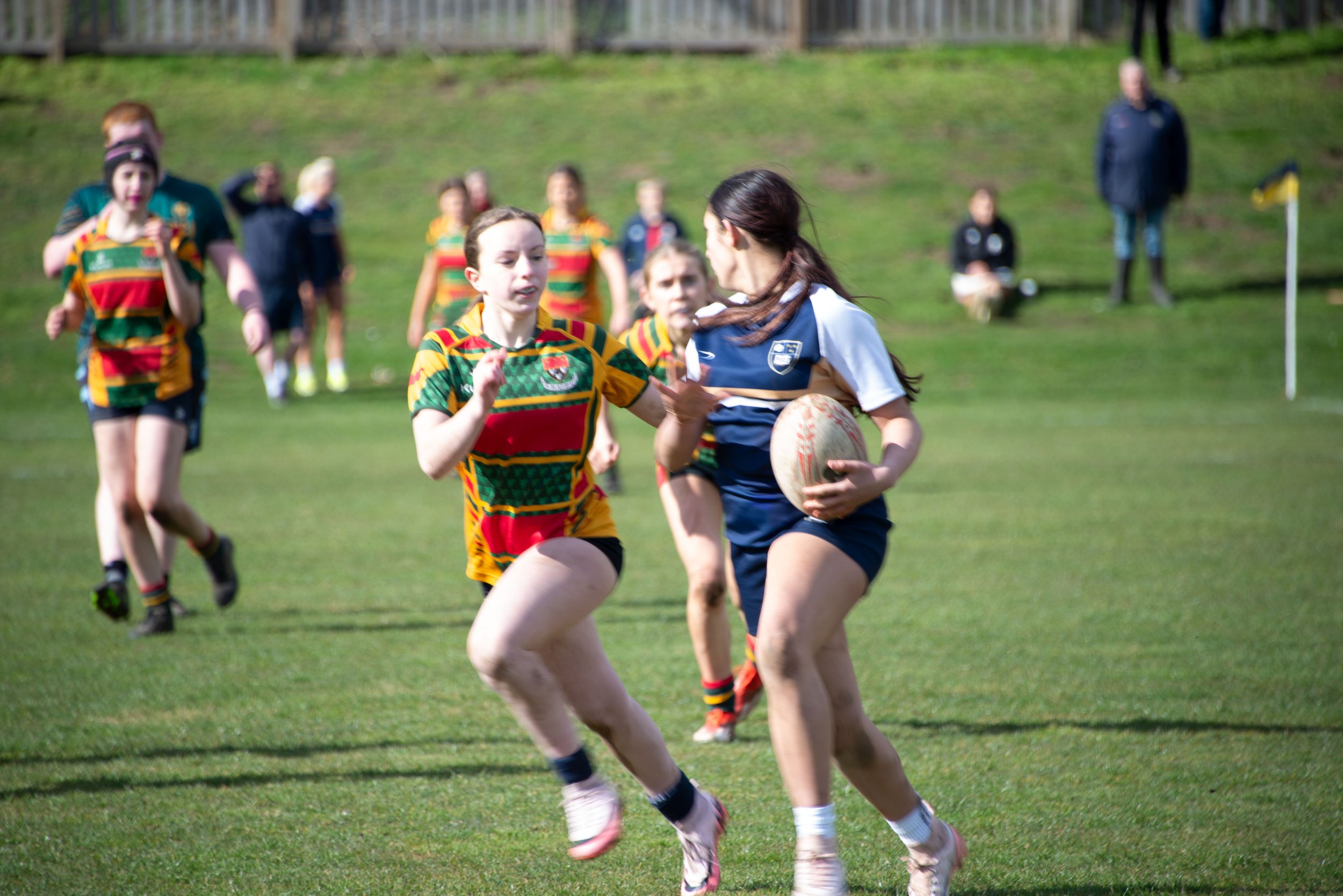 Young girls playing rugby on a grassy field during daytime, wearing colorful sports uniforms with a wooden fence in the background.