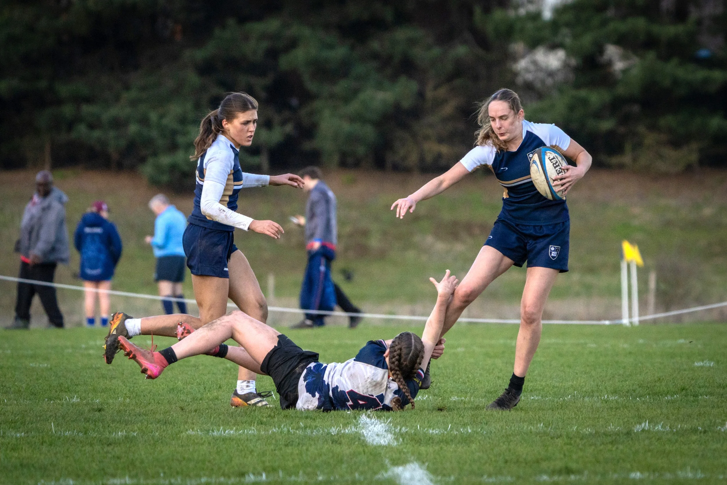 Women playing rugby on a grassy field during daytime, with one woman in a black uniform falling while another in blue and white runs with the ball. In the background, people watch the game, and trees are visible.
