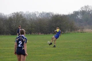 A person wearing a yellow cap and blue shirt is kicking a rugby ball on a grassy field, with three other people watching.