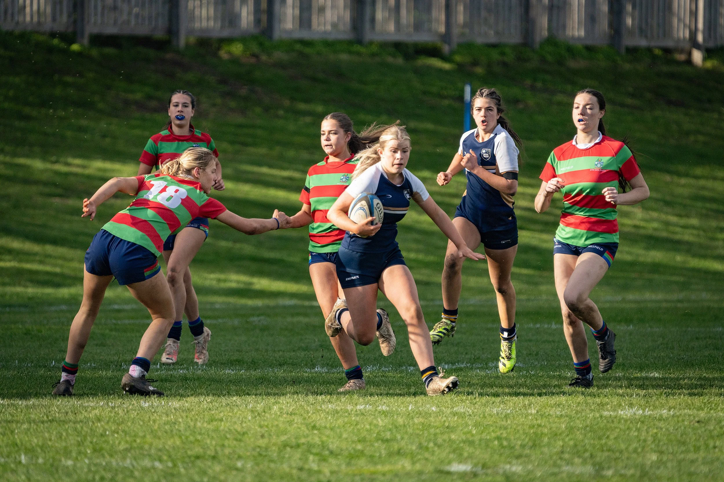Young girls playing rugby on a grassy field, wearing colorful uniforms and carrying a rugby ball.