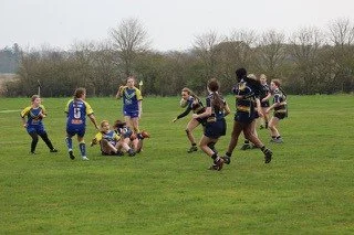 A women's rugby match in progress on a grassy field with players tackling and running, while children watch nearby.