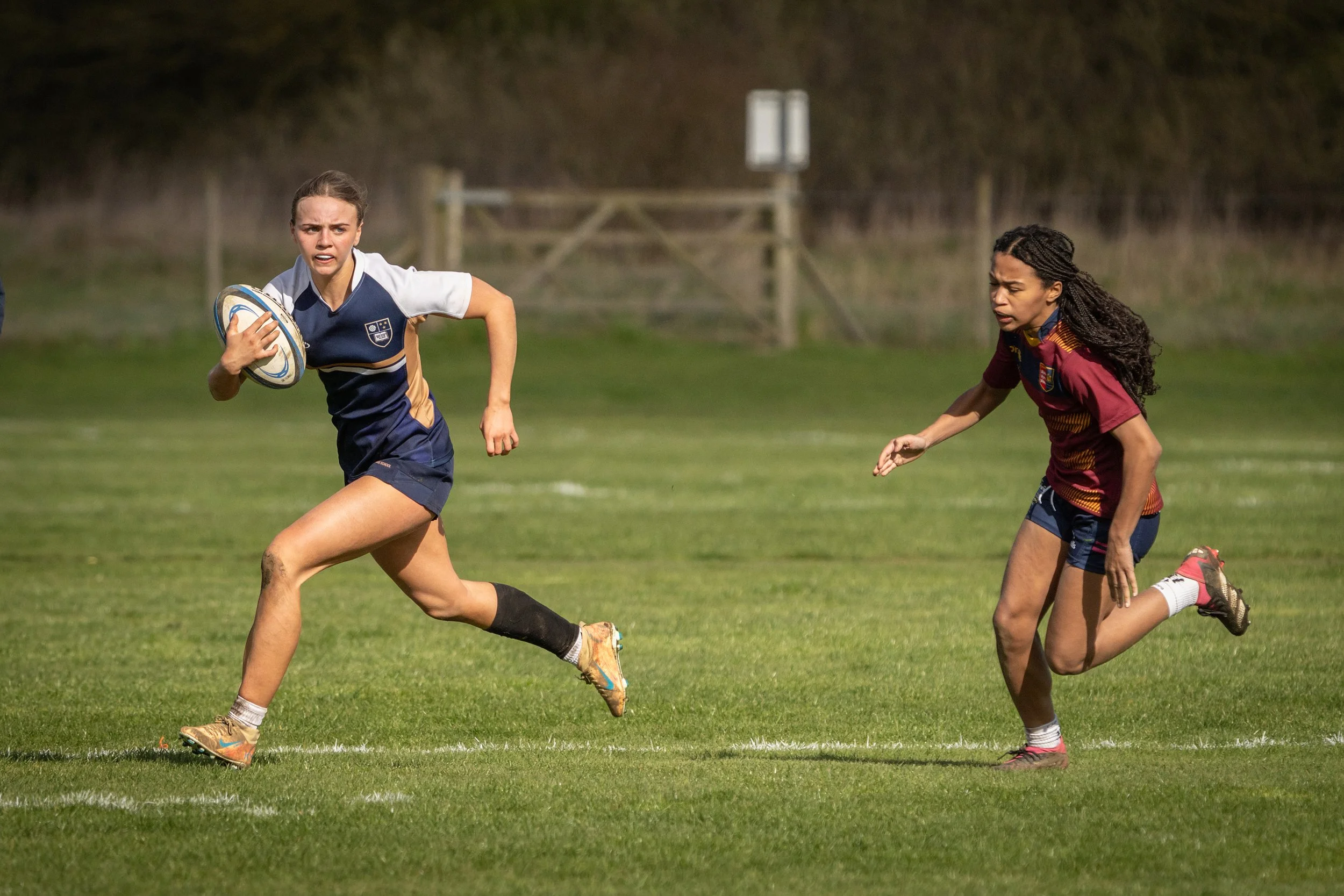 Two female rugby players running on a grassy field during a game, one holding a rugby ball and the other closely behind, both in athletic gear.