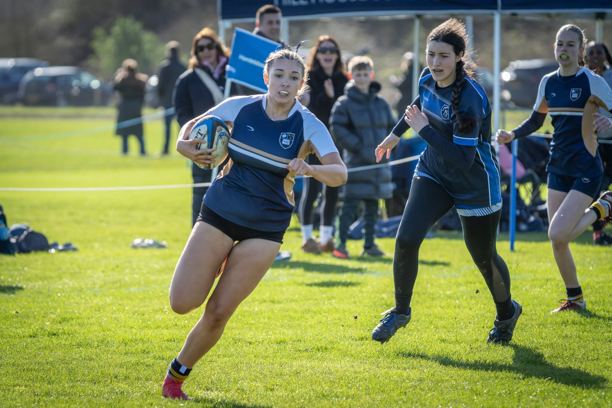 A women's rugby game in progress with a player running holding a rugby ball and other players in pursuit on a grassy field. Spectators are watching in the background.
