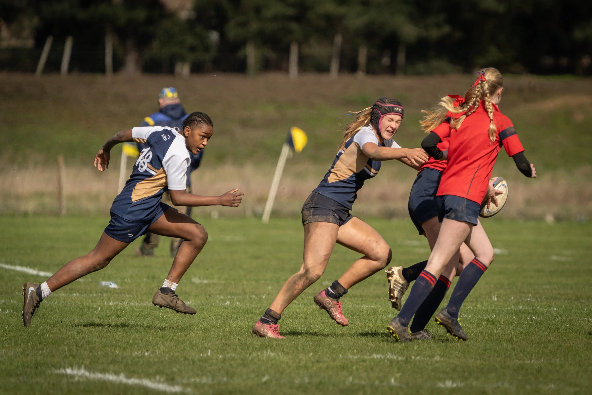 Women playing rugby on a grassy field with trees in the background.