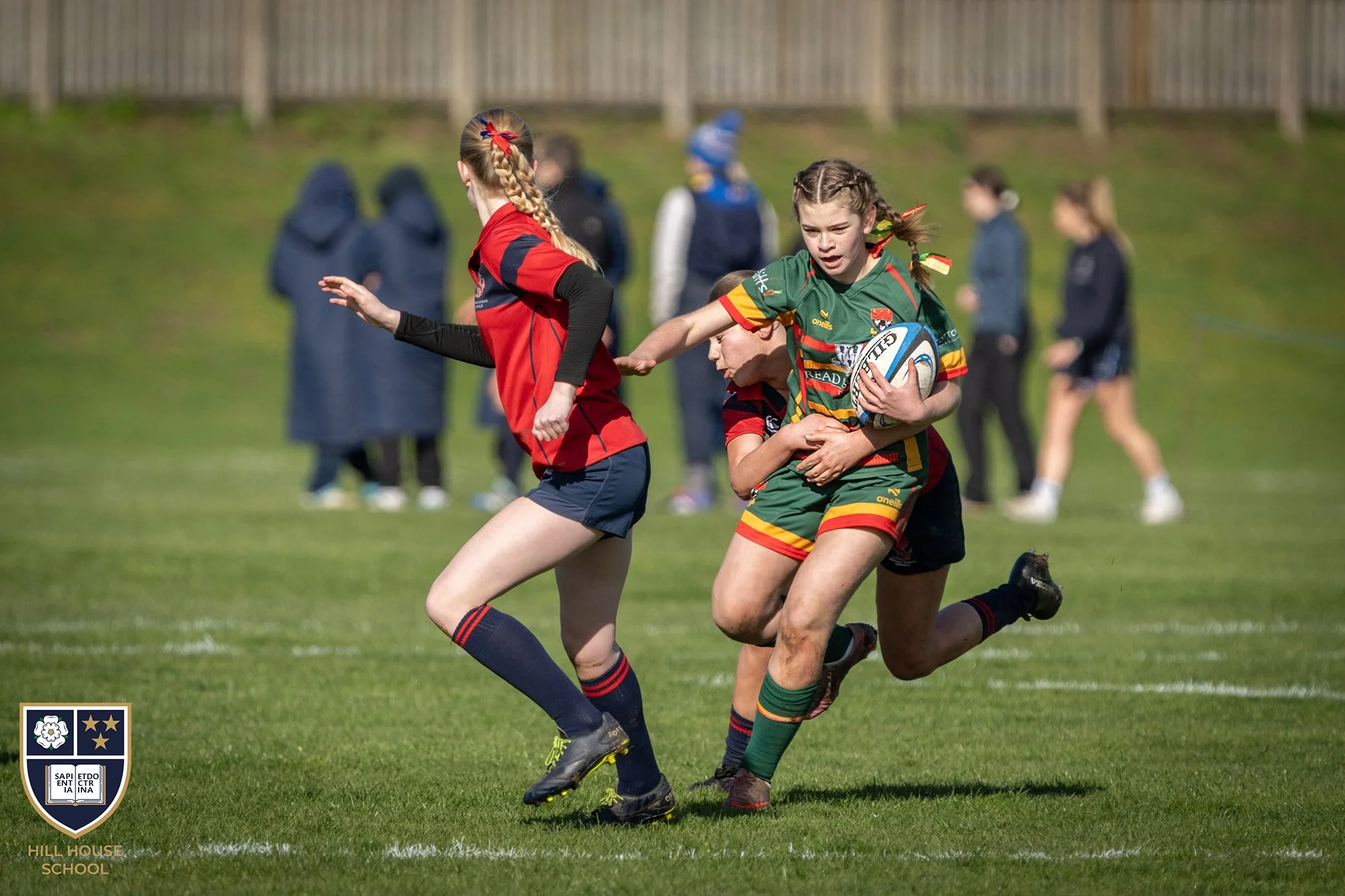 Young girls playing rugby on a grassy field during daytime, dressed in colorful sports uniforms, with spectators and a fence in the background.
