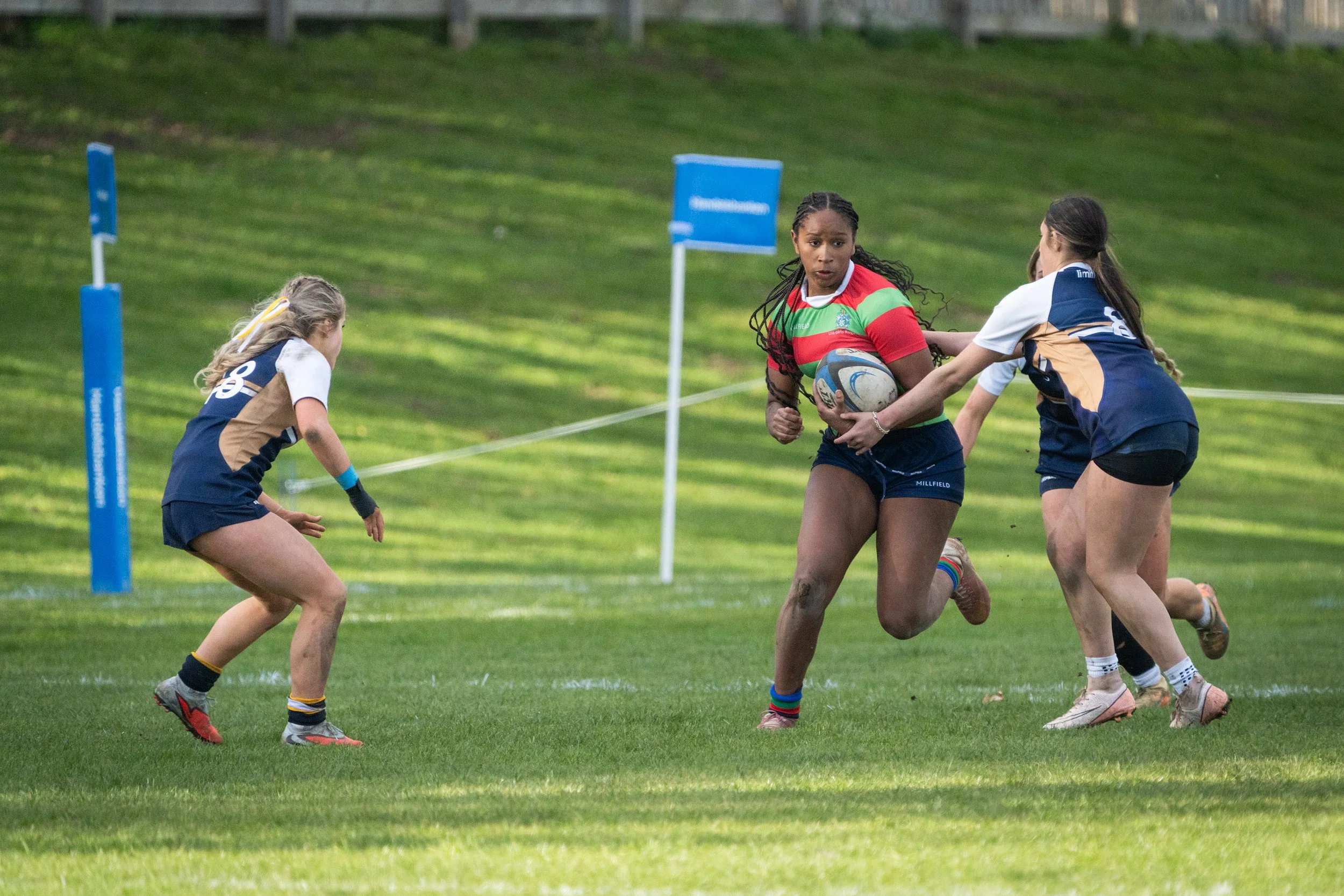 Women playing rugby on a grassy field, with one woman holding a rugby ball and others approaching her.