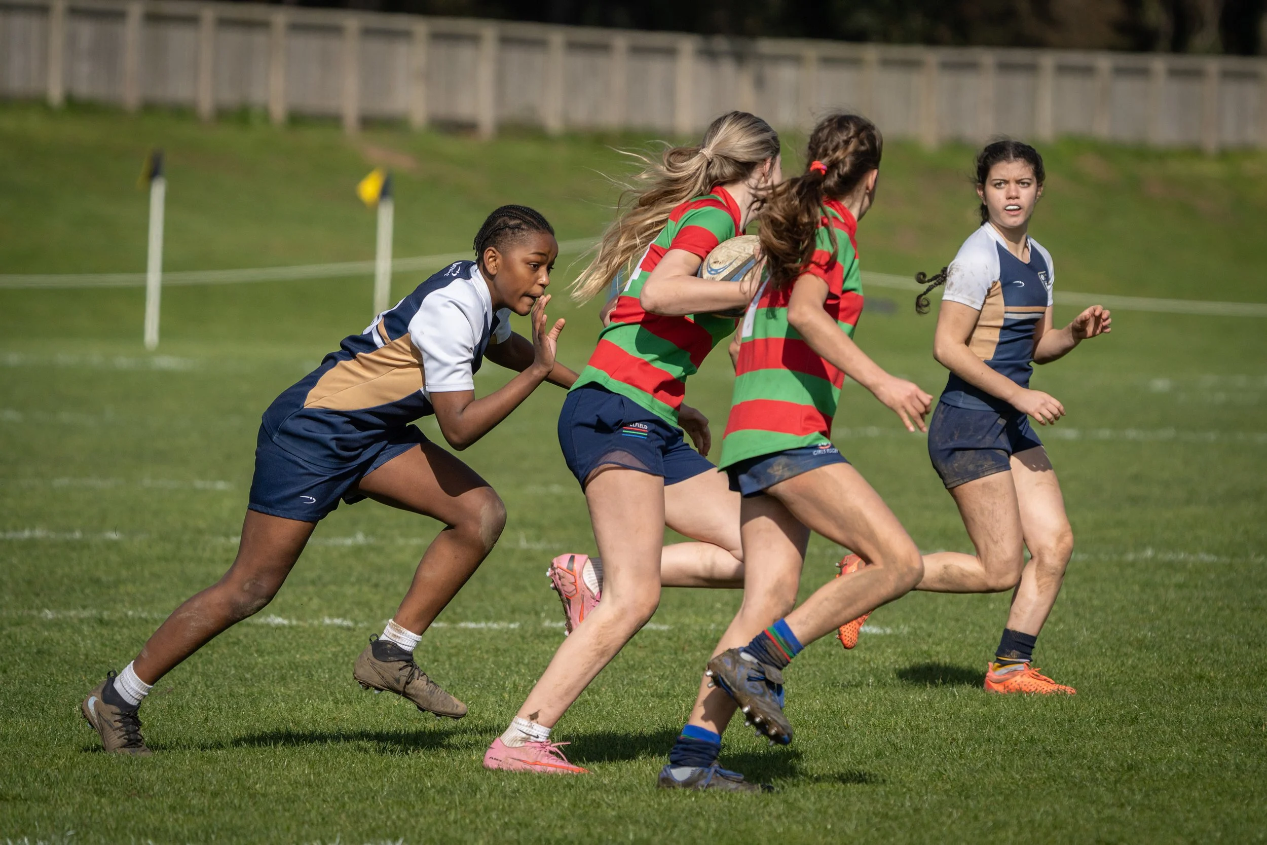 Girls playing rugby on a grassy field. One girl in a red and green striped jersey holds a rugby ball, while others in blue and tan uniforms run alongside.