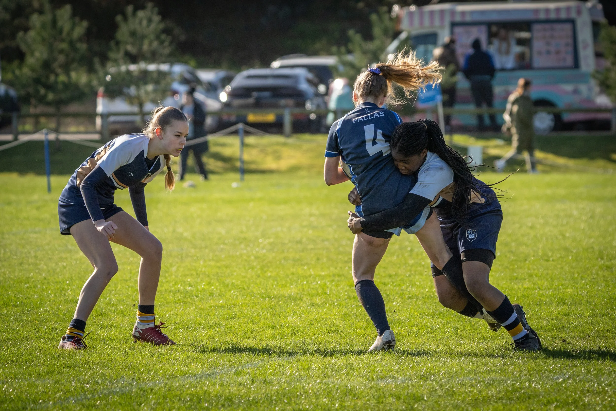 Two women in sports uniforms playing rugby on a grassy field, with one holding the other in a tackle, and a third woman watching nearby.