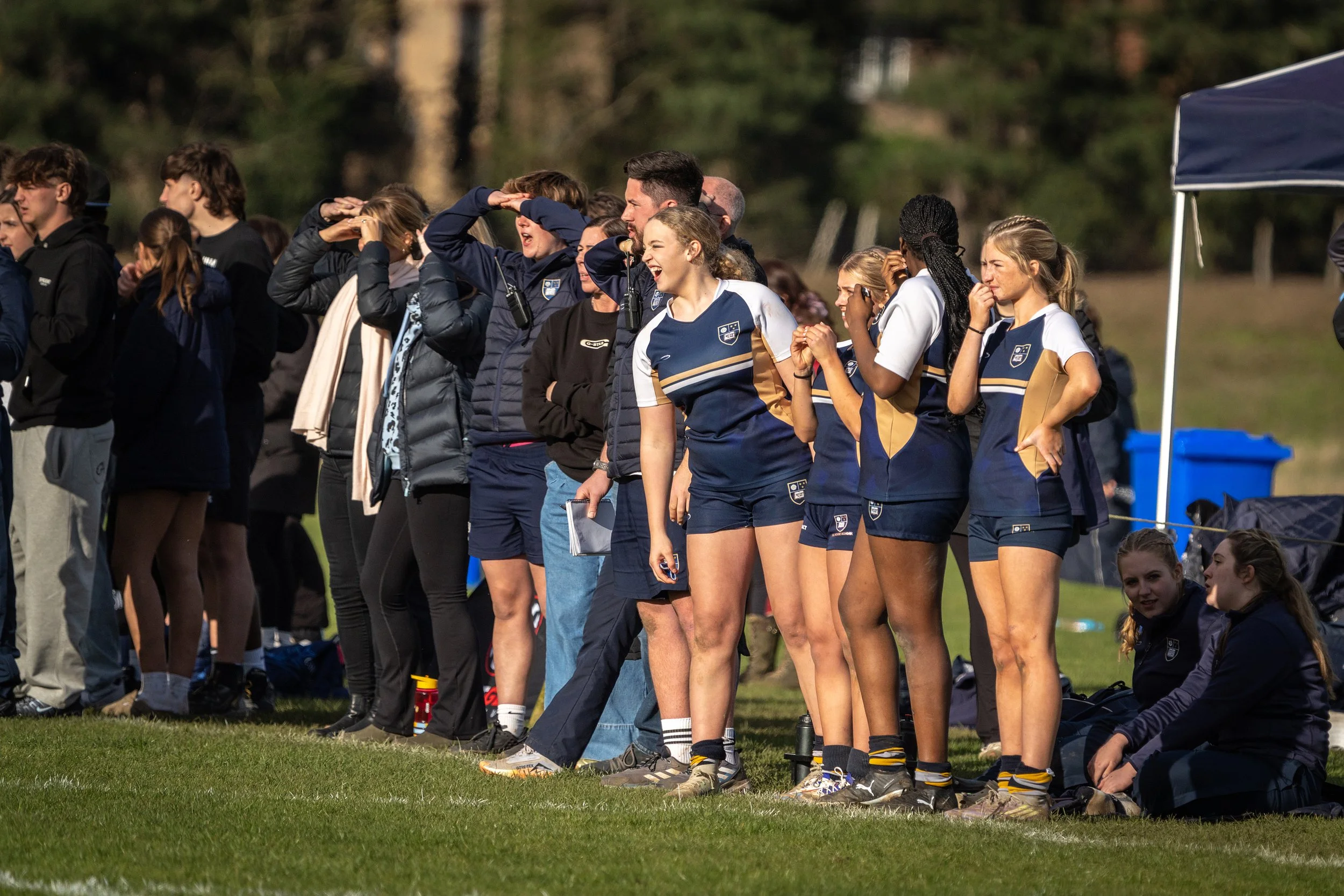Female rugby players and spectators standing on the sideline during a game, some shielding their eyes from the sun.