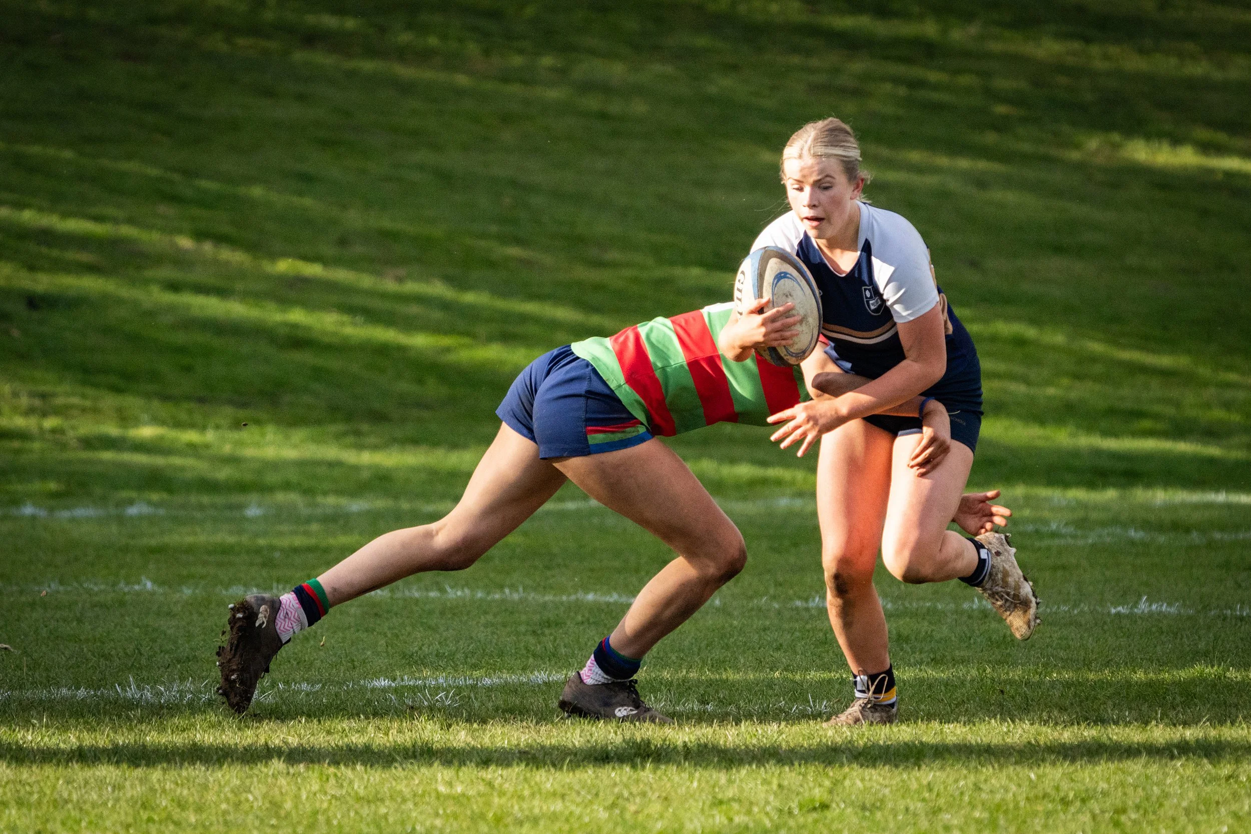 A female rugby player with blonde hair in a ponytail tackling an opponent in a green and red striped jersey on a grassy field during a rugby match.