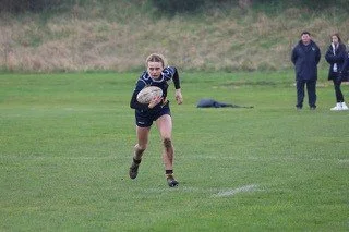 A girl running on a grassy sports field holding a rugby ball, with two people watching in the background.