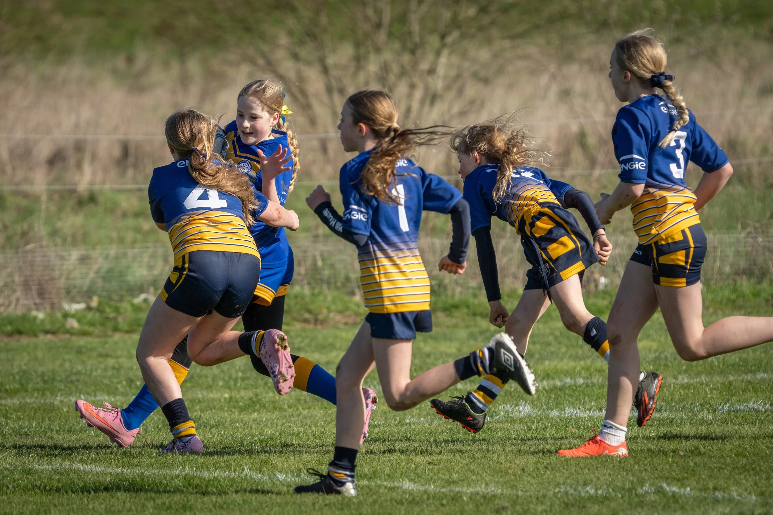 Young girls playing rugby outdoors on grass field, wearing blue and yellow sports uniforms.