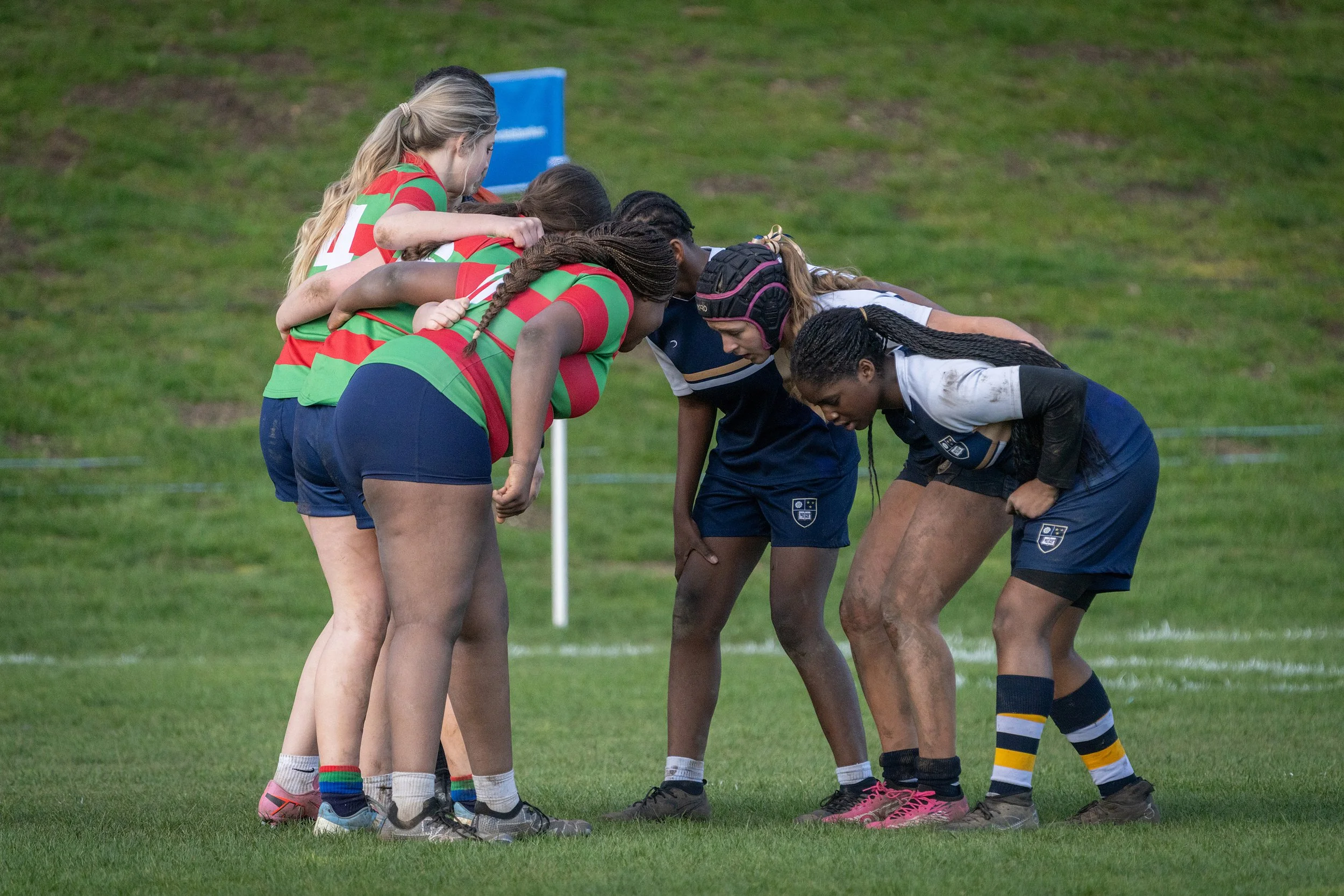 Rugby players on the field, forming a scrum, with two teams of young women wearing team uniforms and protective gear.