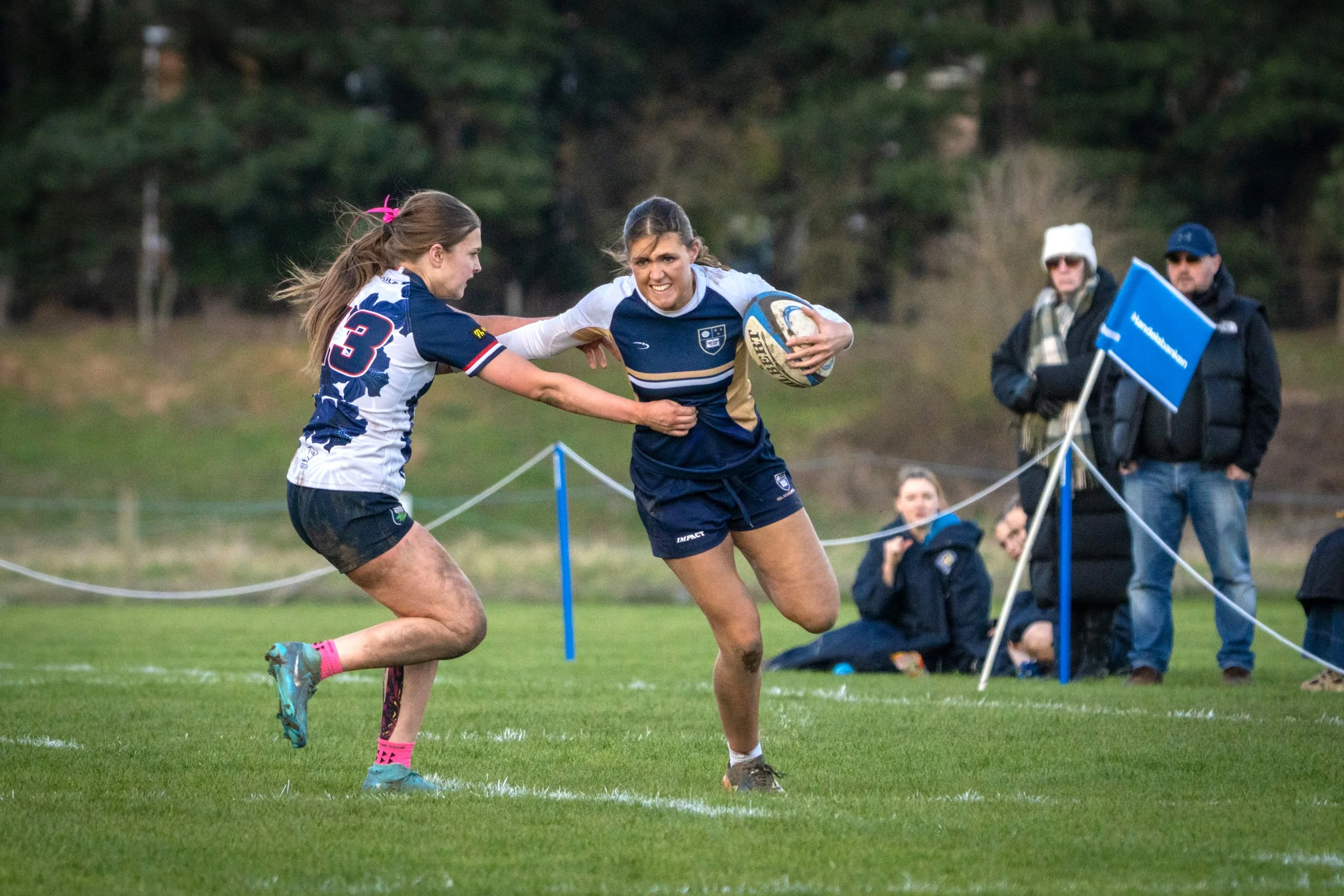 Two female rugby players are competing for the ball during a match on a grassy field, with spectators and trees in the background.