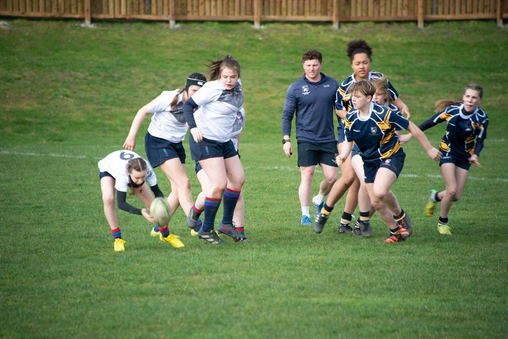 Young female rugby players in action on the field, competing for possession of the ball with coaches observing.