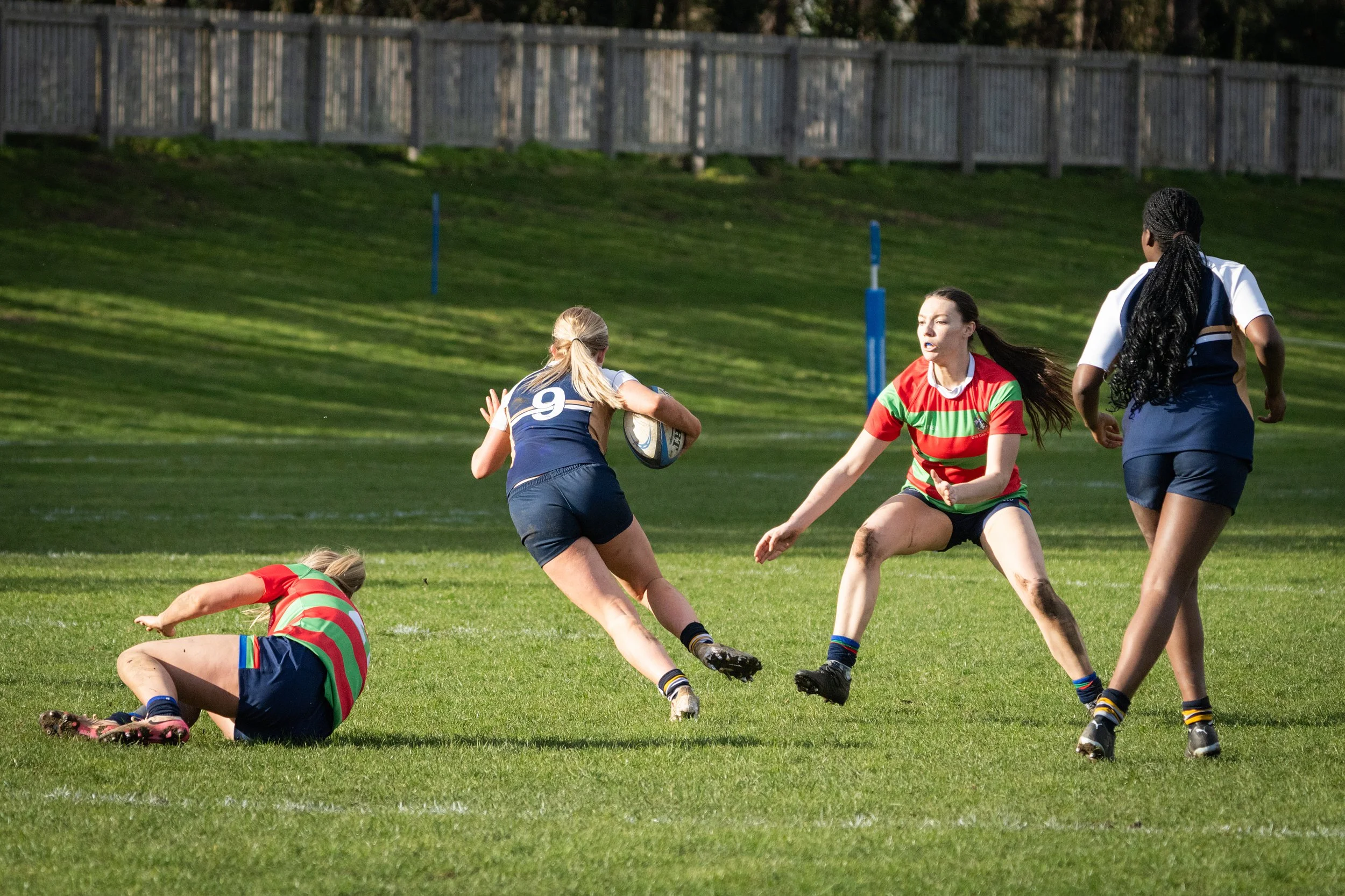 Women playing rugby on a grassy field during daytime, wearing colorful uniforms. One woman in a red and green striped jersey is being tackled by another woman in a blue and white uniform, while others look on nearby.