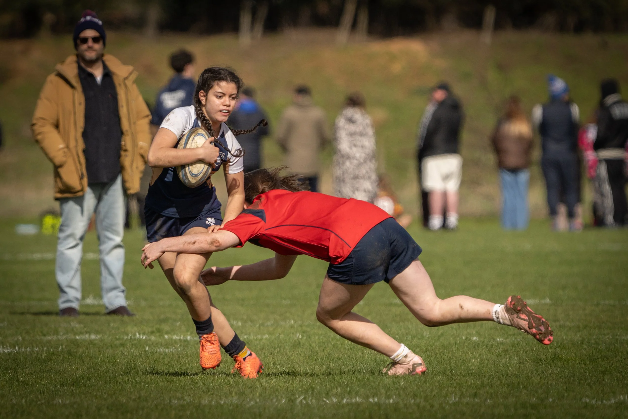 A rugby match in progress with two female players on the ground, one in red and the other in blue, grabbing the ball, while a male referee or coach stands nearby. Several spectators stand in the background on a grassy field.