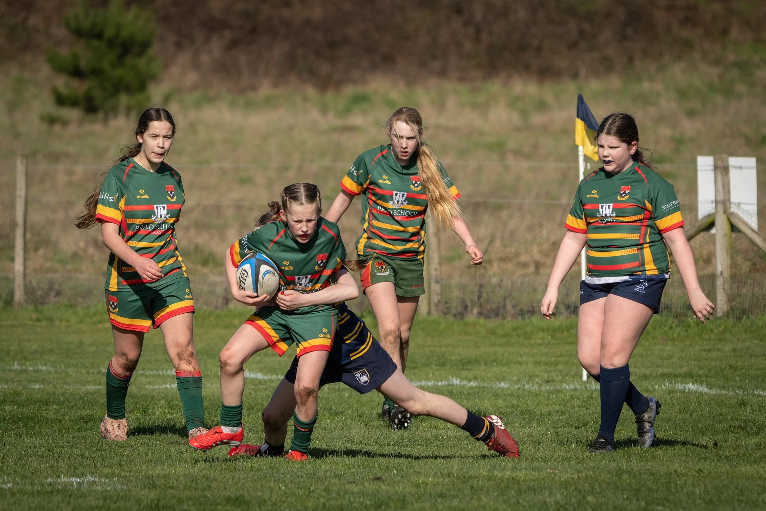 Young girls playing rugby on a grassy field, wearing green and red uniforms.