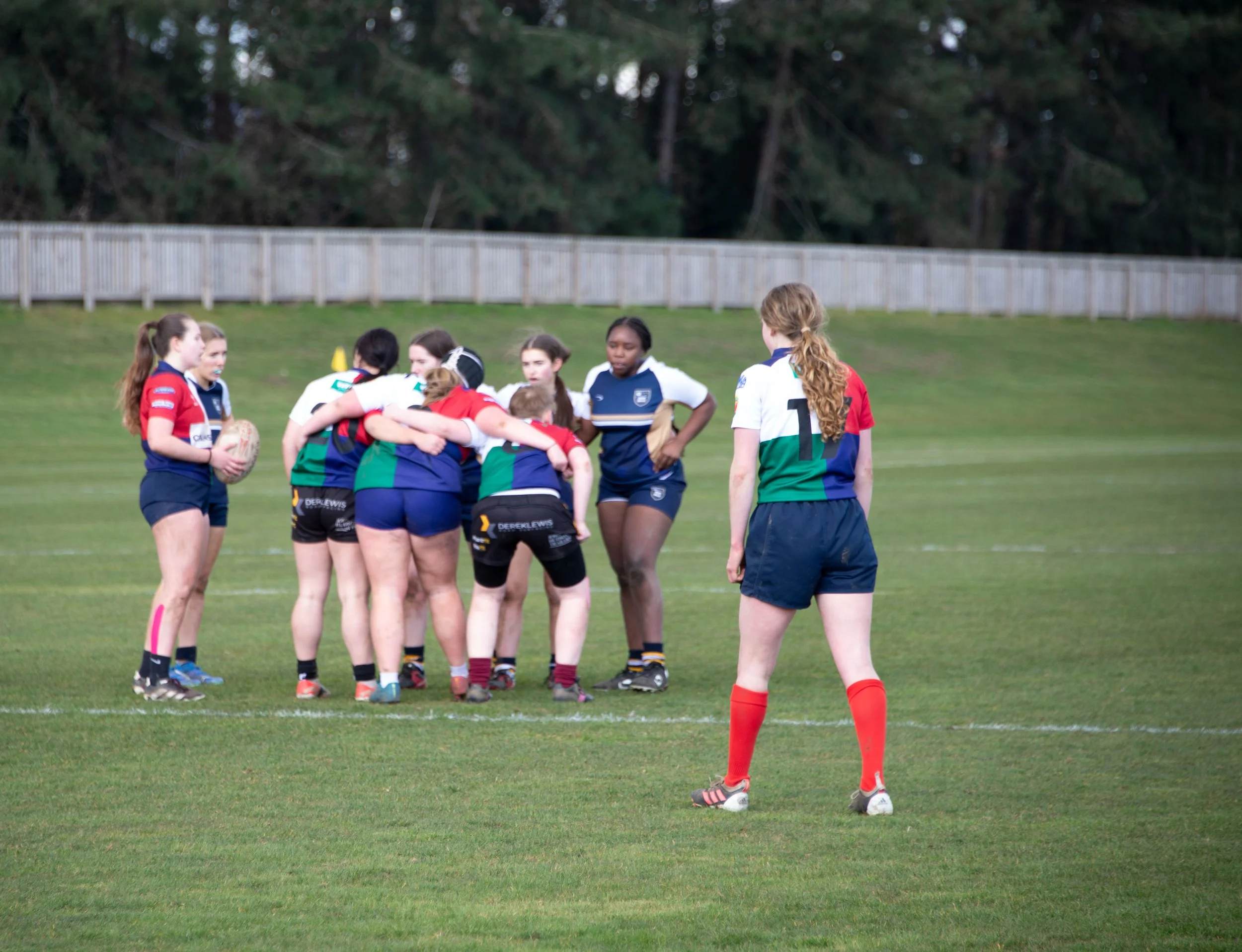 A group of female rugby players on a field, some huddling, with a referee standing nearby, surrounded by trees and a wooden fence in the background.