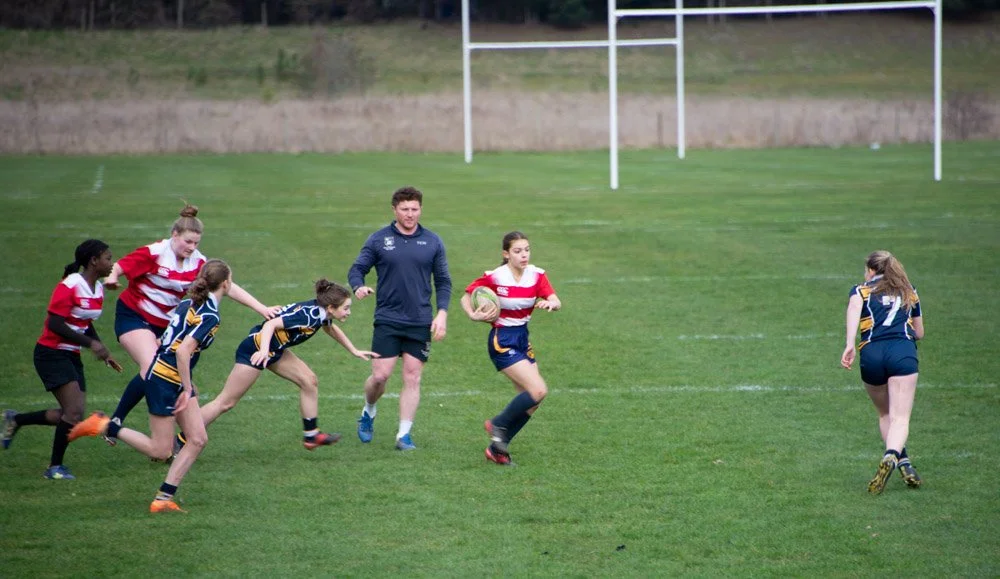 Women’s rugby game on a green field with players running and a referee in the background.